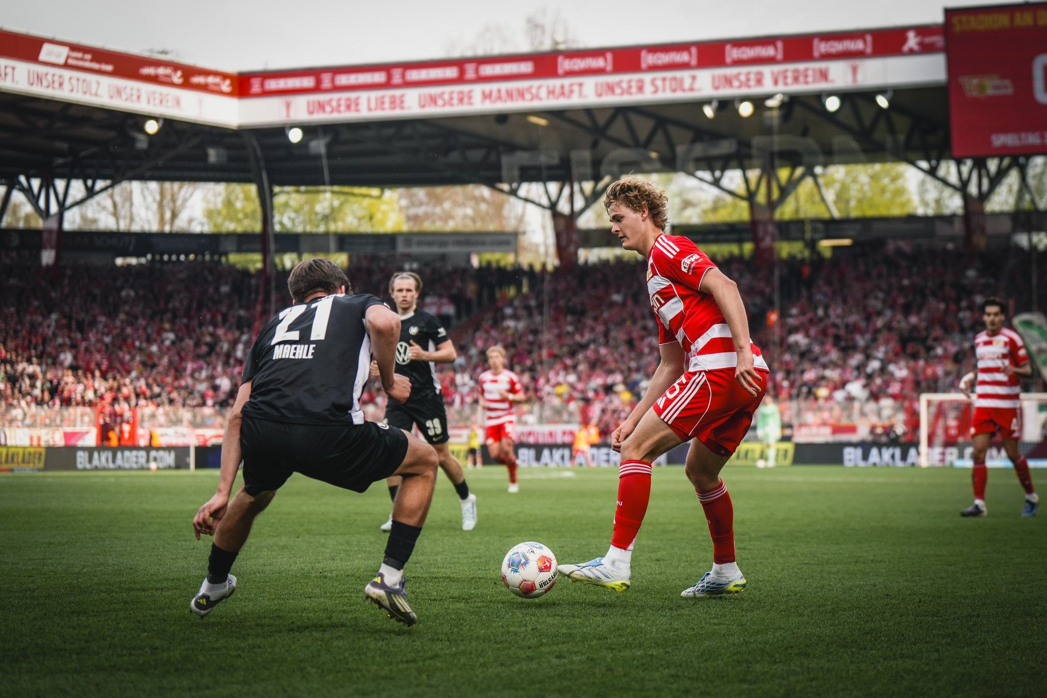 Fußballspiel auf einem Stadionfeld, Spieler in roten und weißen Trikots dribbelt den Ball, gegnerischer Spieler versucht, ihn zu stoppen.