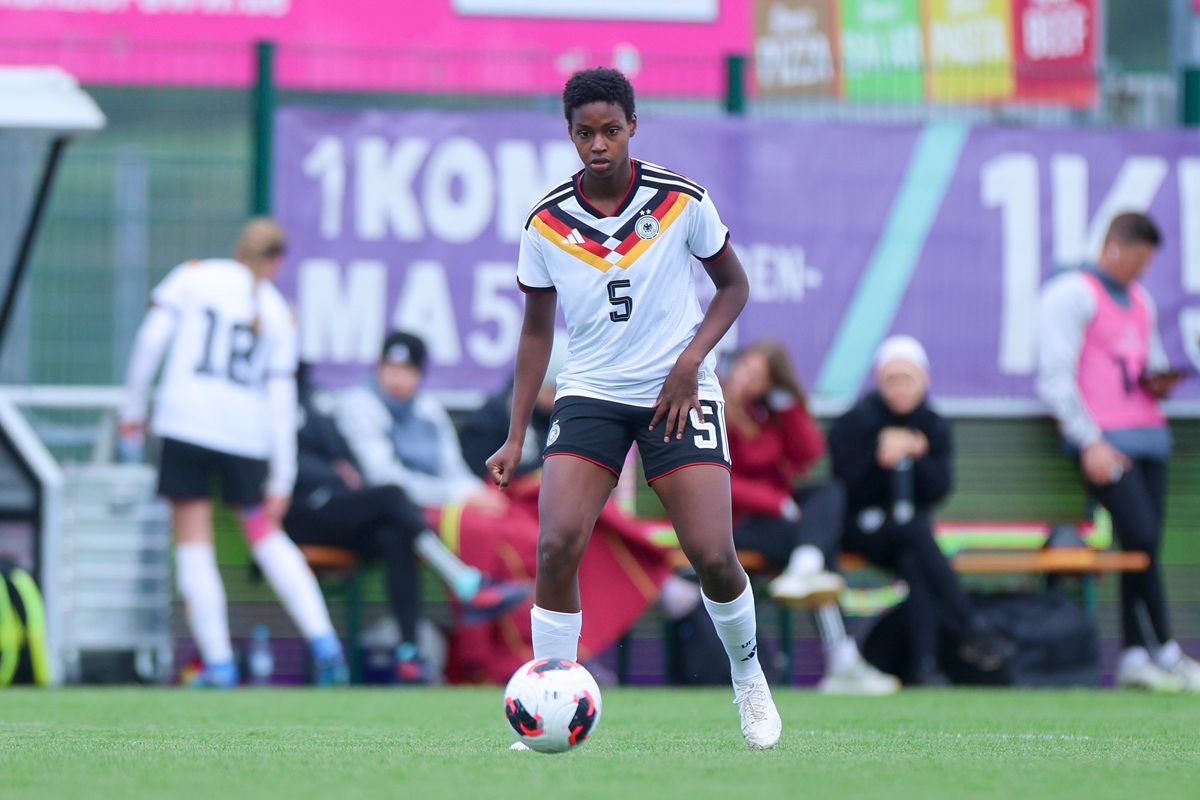 Eine Spielerin in einem weißen Fußballtrikot mit deutschen Farben dribbelt den Ball auf dem Spielfeld.