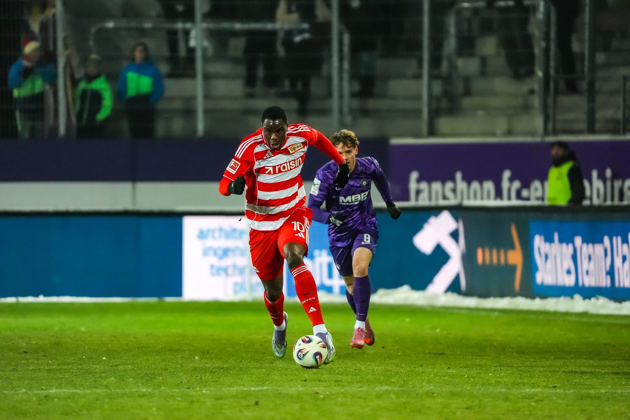 Ein Fußballspieler in rotem Trikot rennt mit dem Ball, während ein Verteidiger in lila versucht, ihn einzuholen.