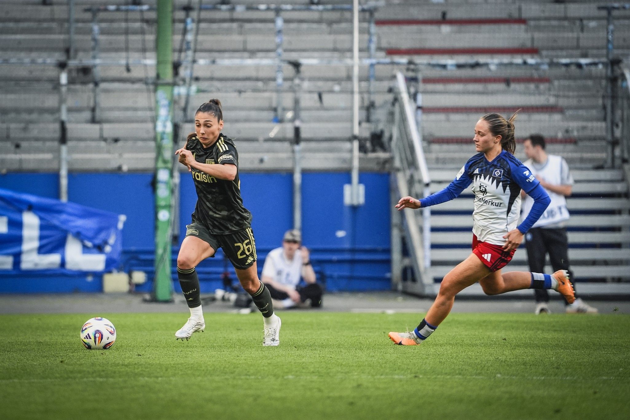 Zwei Fußballspielerinnen laufen mit dem Ball über das Spielfeld, eine in einem schwarzen Trikot, die andere in einem weißen und blauen Trikot.