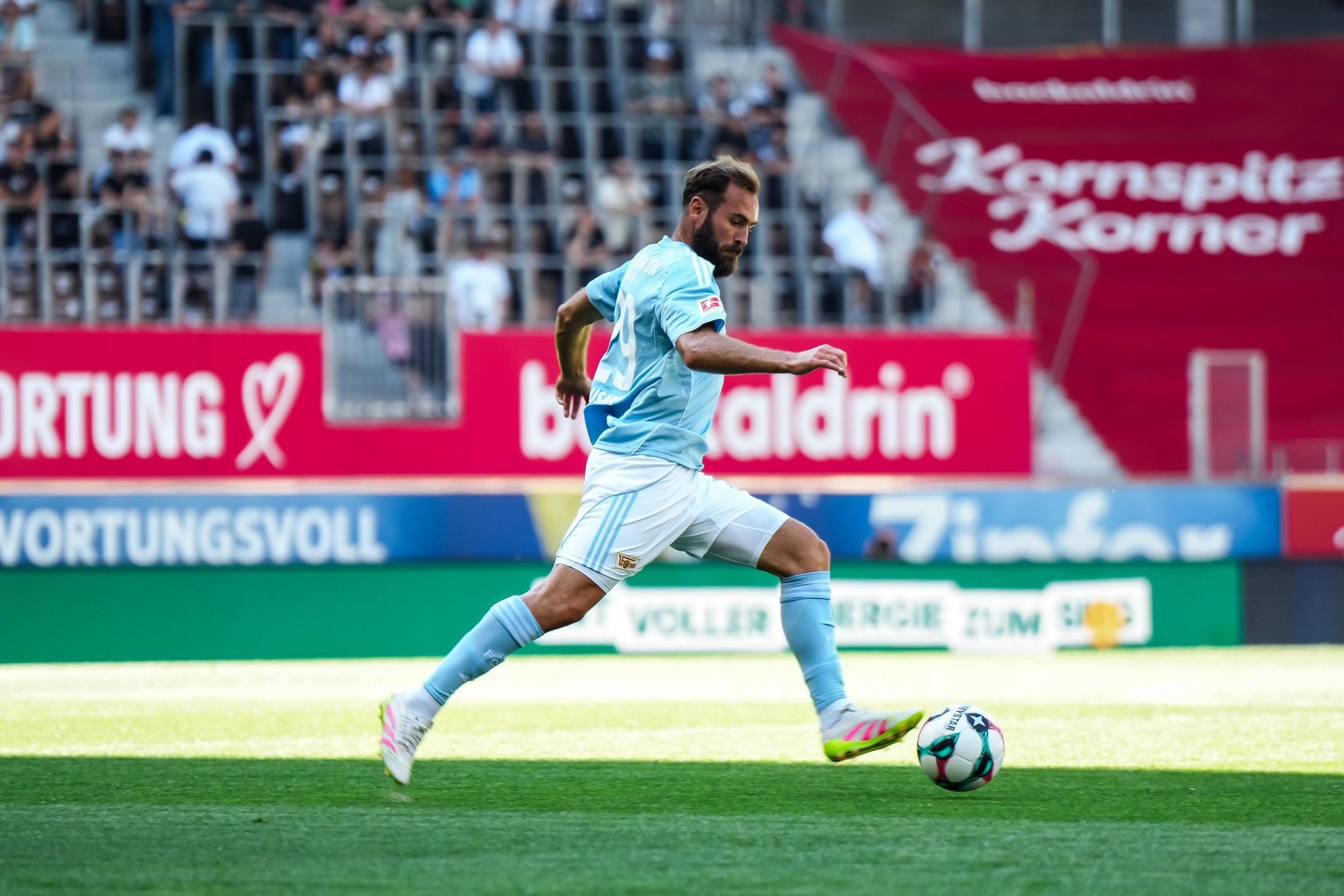 Ein Fußballspieler in hellblauem Trikot sprintet über das Spielfeld. Zuschauer im Hintergrund sind zu sehen.