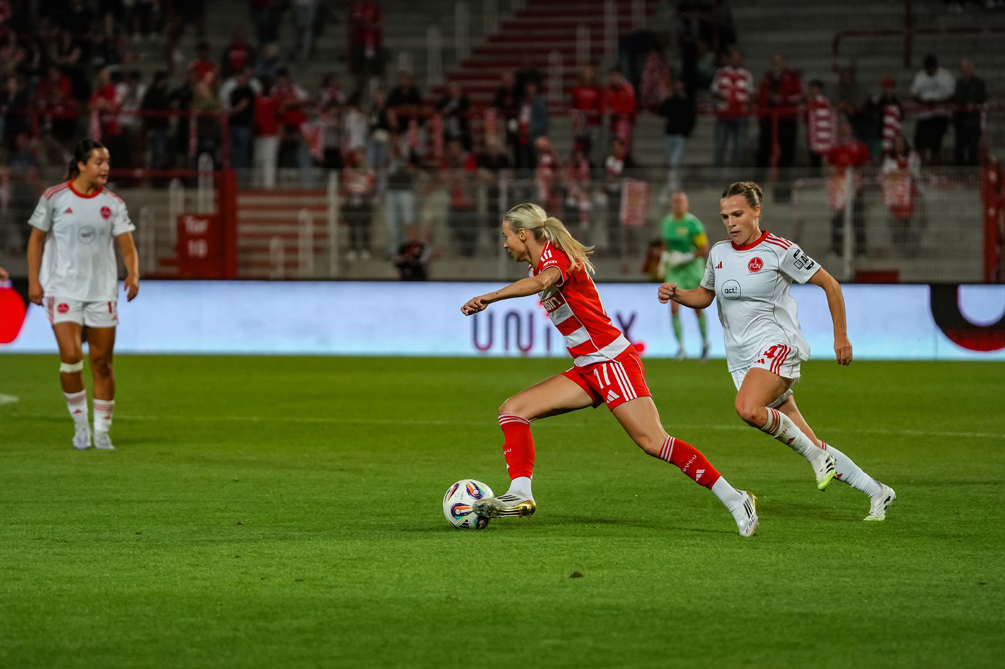 Eine Fußballspielerin in rotem Trikot dribbelt den Ball, während eine Gegnerin in weißem Trikot sie verfolgt. Zuschauer im Hintergrund.
