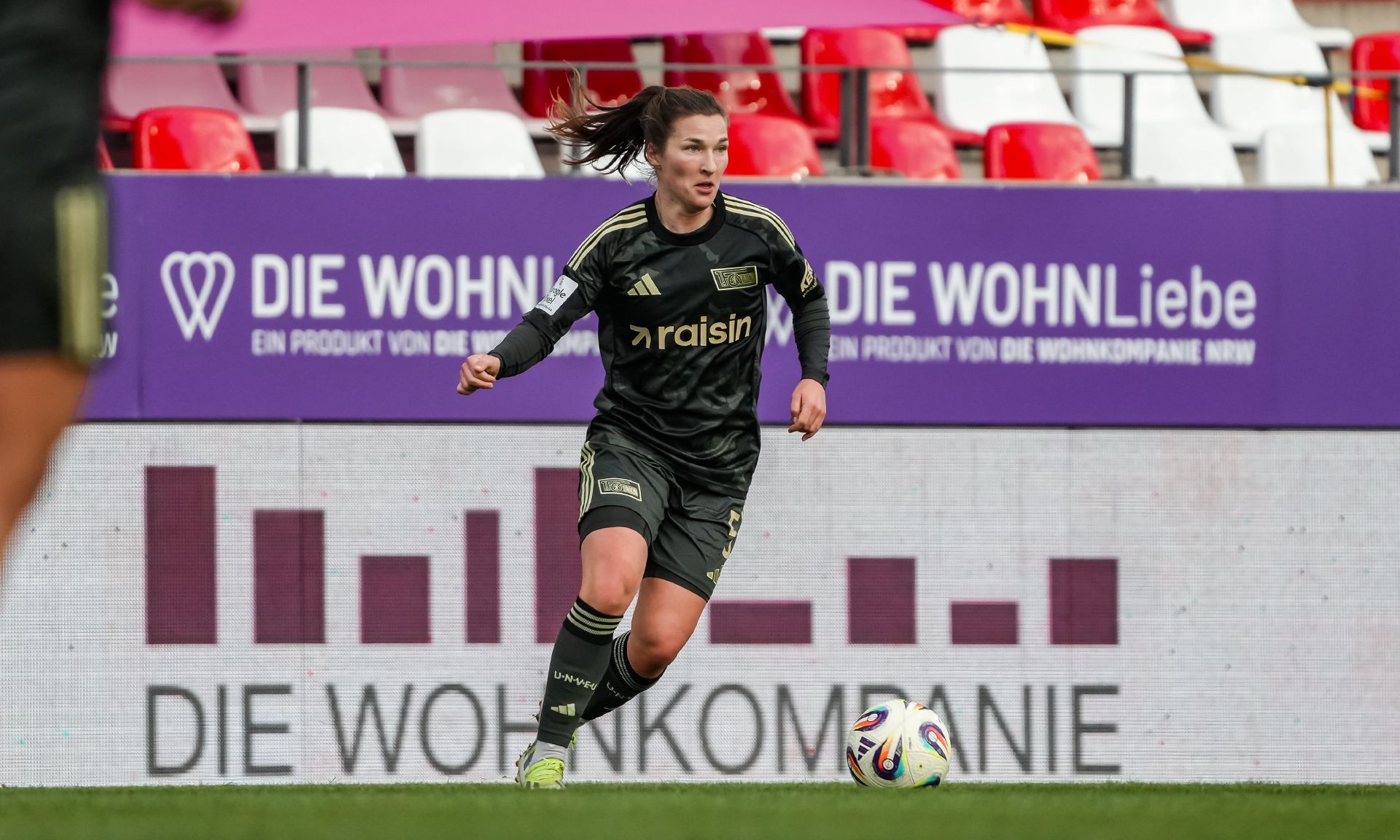 Spielerin in schwarzer Fußballausrüstung mit "raisin"-Logo läuft auf dem Spielfeld, im Hintergrund Werbebanner.