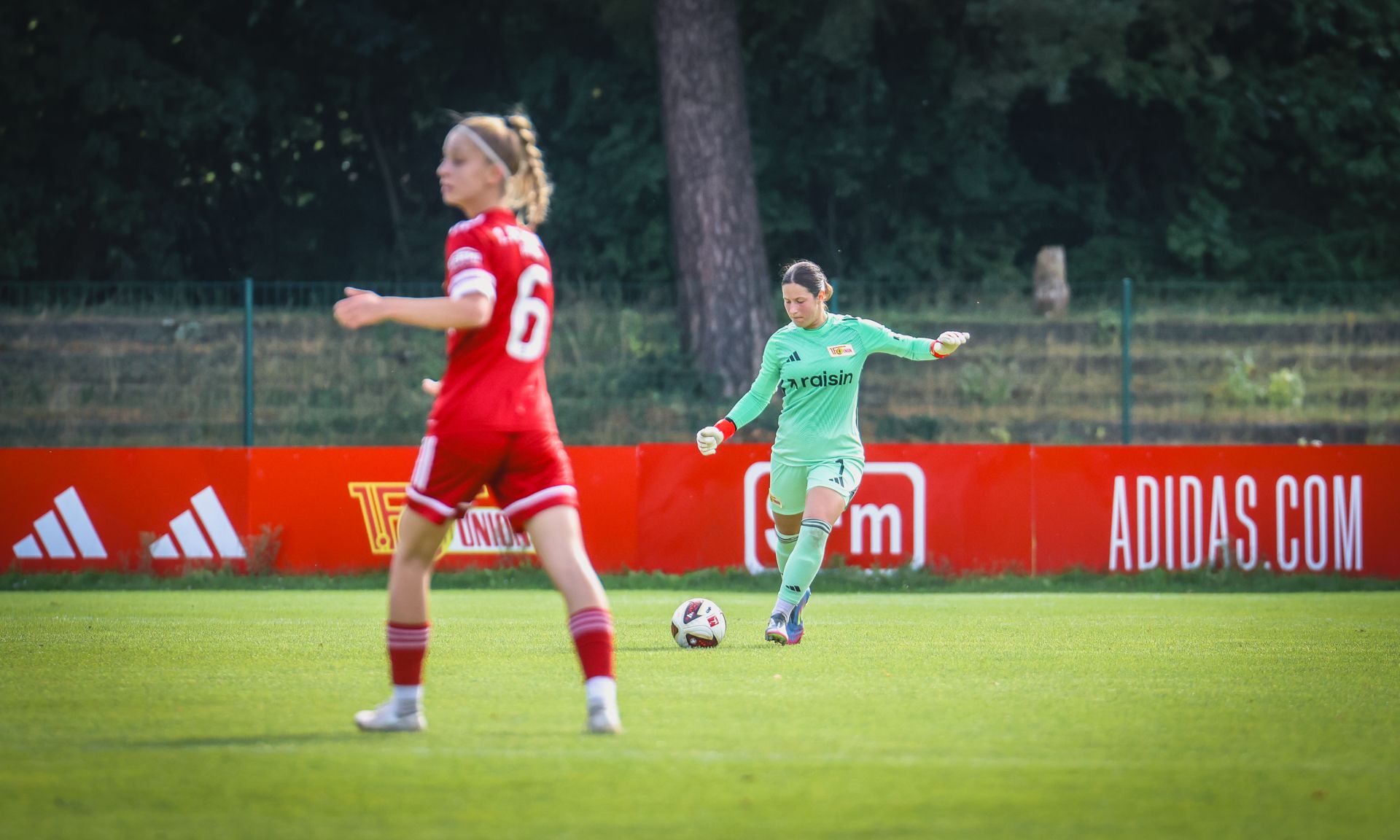 Torhüterin in grün schlägt den Ball auf einem Fußballfeld, während eine Spielerin in rot in der Nähe positioniert ist.