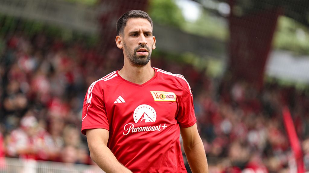 Ein Fußballspieler in rotem Trikot mit einem Sponsorenlogo, der auf dem Spielfeld steht. Im Hintergrund eine Menschenmenge.
