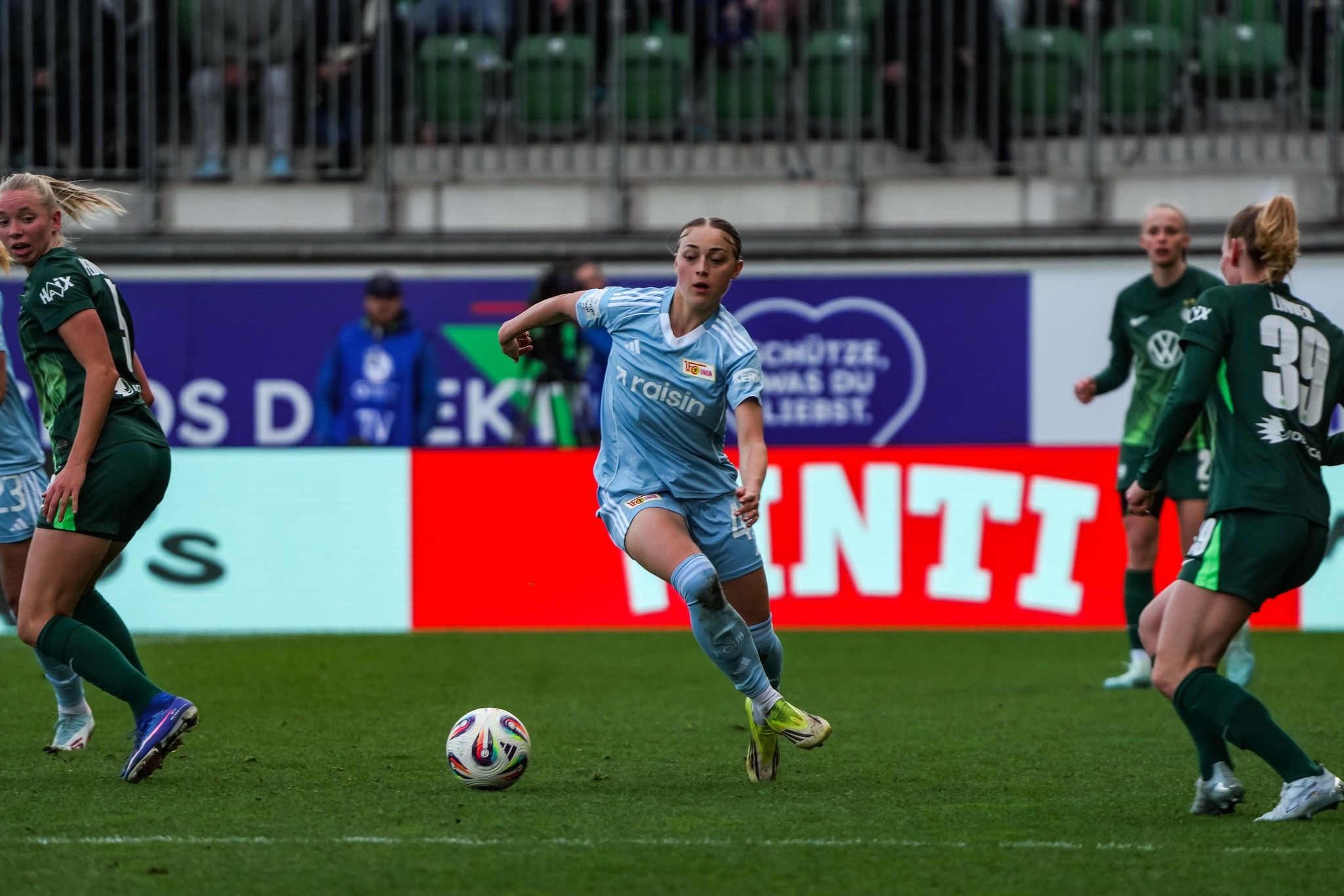 Spielerin in blauem Trikot dribbelt den Ball, umgeben von gegnerischen Spielerinnen in grün. Stadionatmosphäre im Hintergrund.