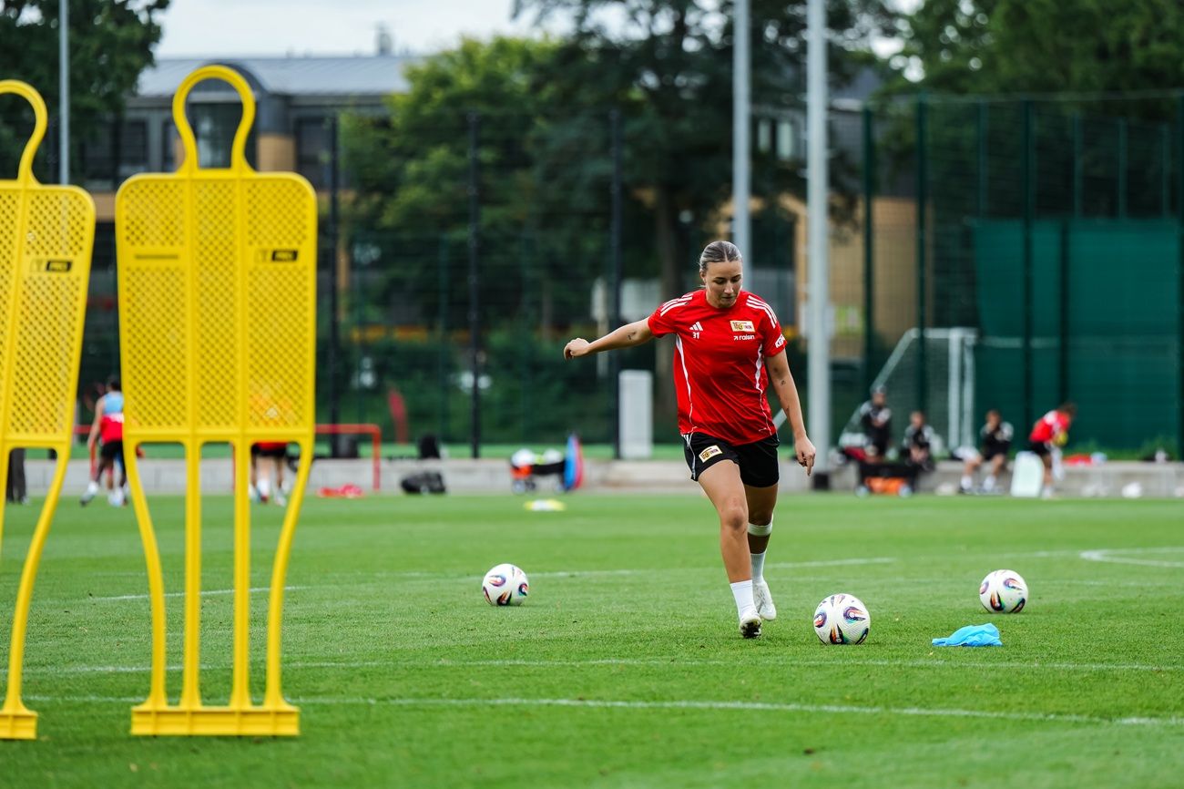 Frau in rotem Trikot dribbelt einen Fußball auf einem Trainingsplatz, mit Hütchen im Hintergrund.