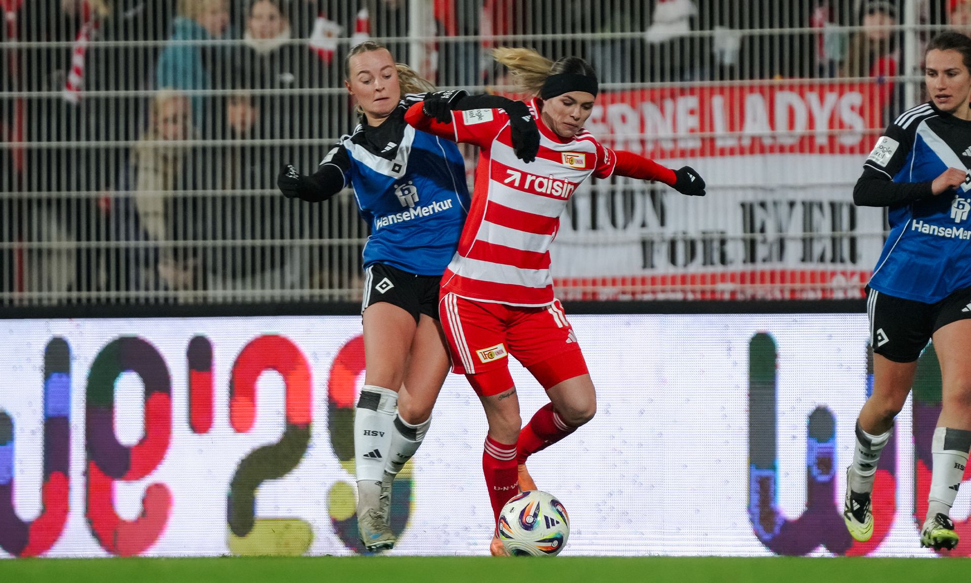 Spielszene im Frauenfußball: Eine Spielerin mit rotem Trikot wird von einer gegnerischen Spielerin mit blauem Trikot umgestoßen.