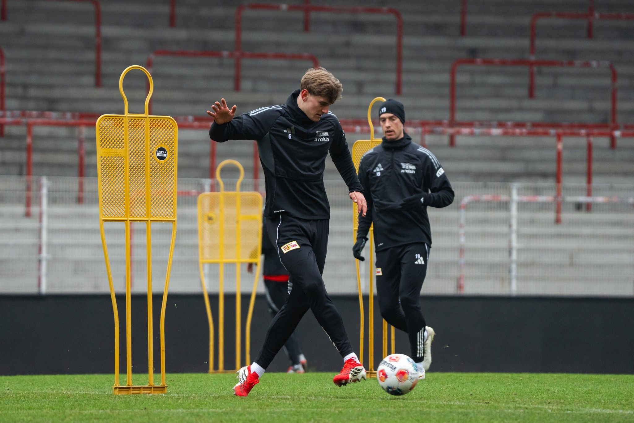 Ein Fussballspieler dribbelt den Ball bei einem Training, während ein Trainer im Hintergrund zuschaut. Gelbe Trainingshilfen stehen auf dem Platz.