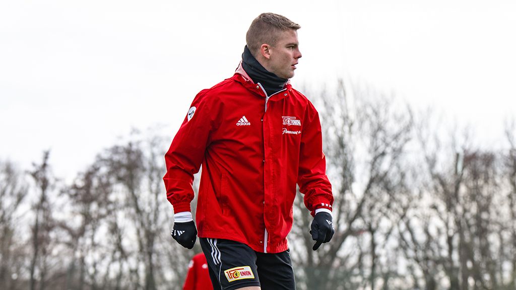 Ein Fußballspieler in roter Jacke und Handschuhen trainiert auf dem Platz, umgeben von Bäumen im Hintergrund.