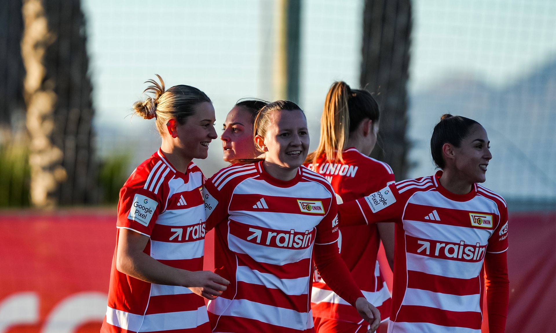 Lächelnde Fußballspielerinnen in roten und weißen Trikots feiern zusammen auf dem Platz.