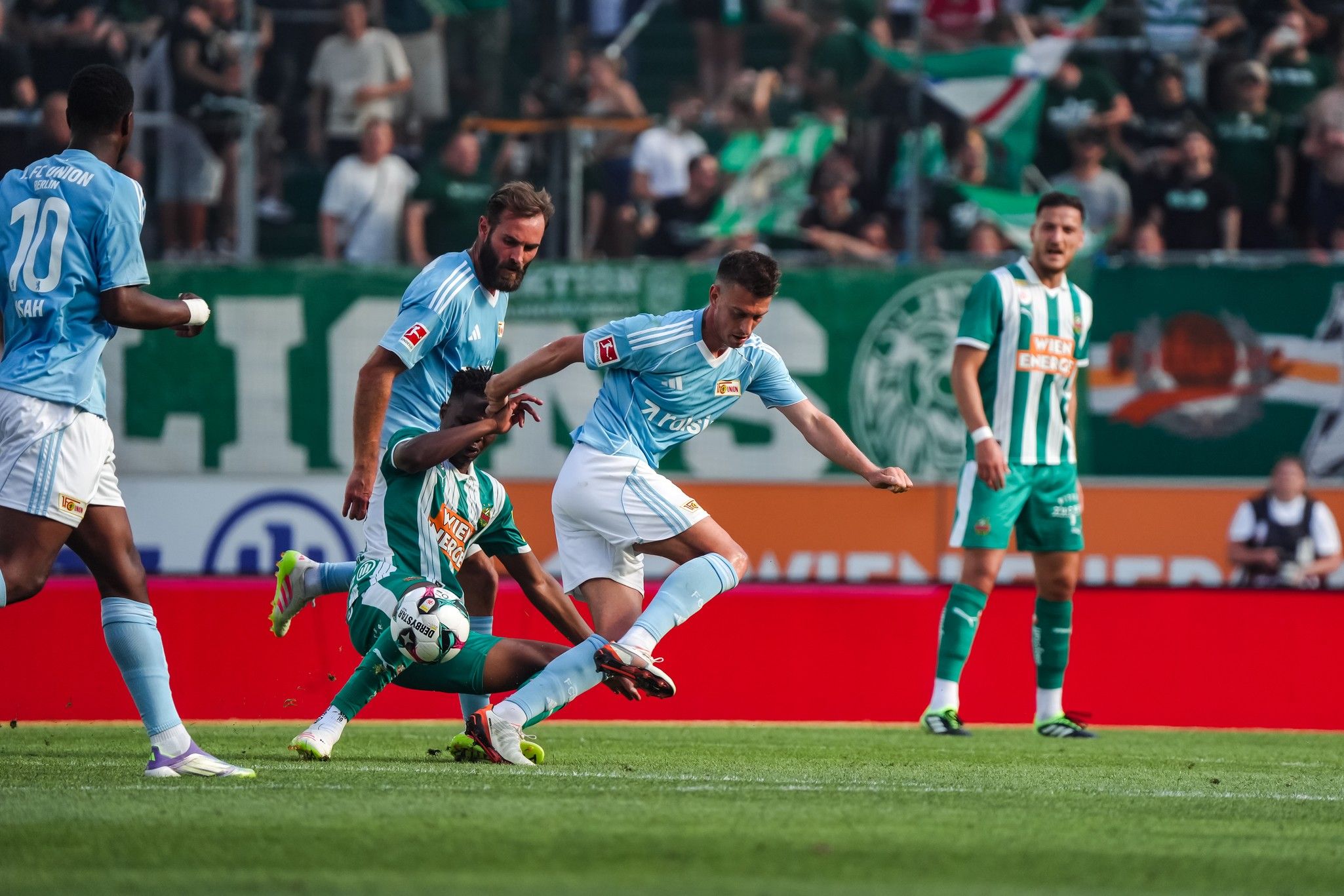 Ein Fußballspieler in blauer Kleidung fällt, während andere Spieler in grüner Kleidung reagieren, auf einem Stadionfeld.