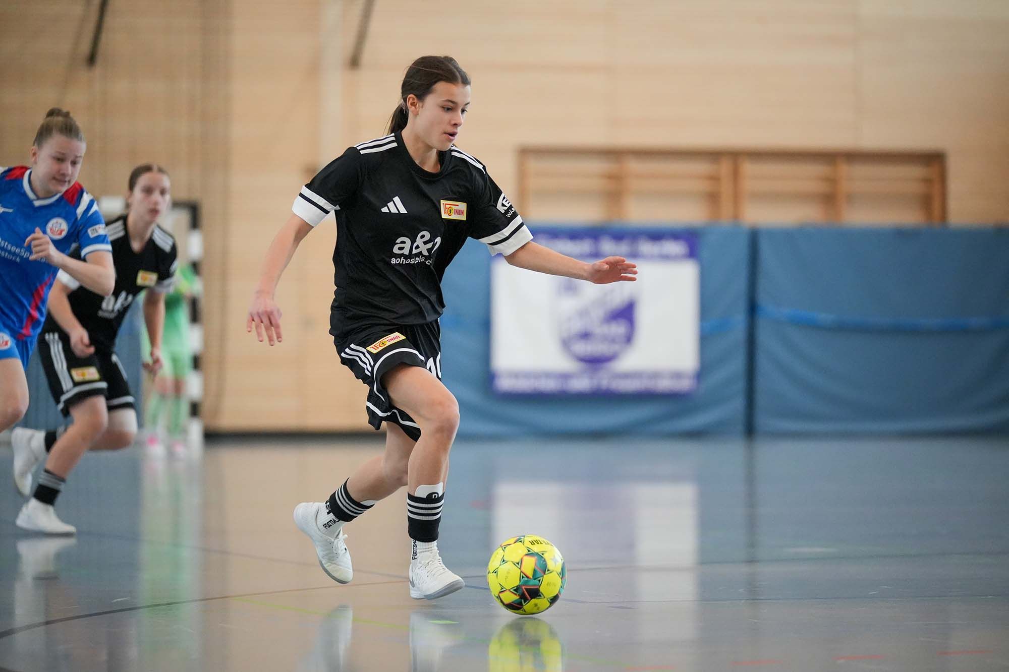 Ein Mädchen in schwarzem Fußballtrikot dribbelt den Ball in einer Halle, während andere Spielerinnen im Hintergrund zu sehen sind.