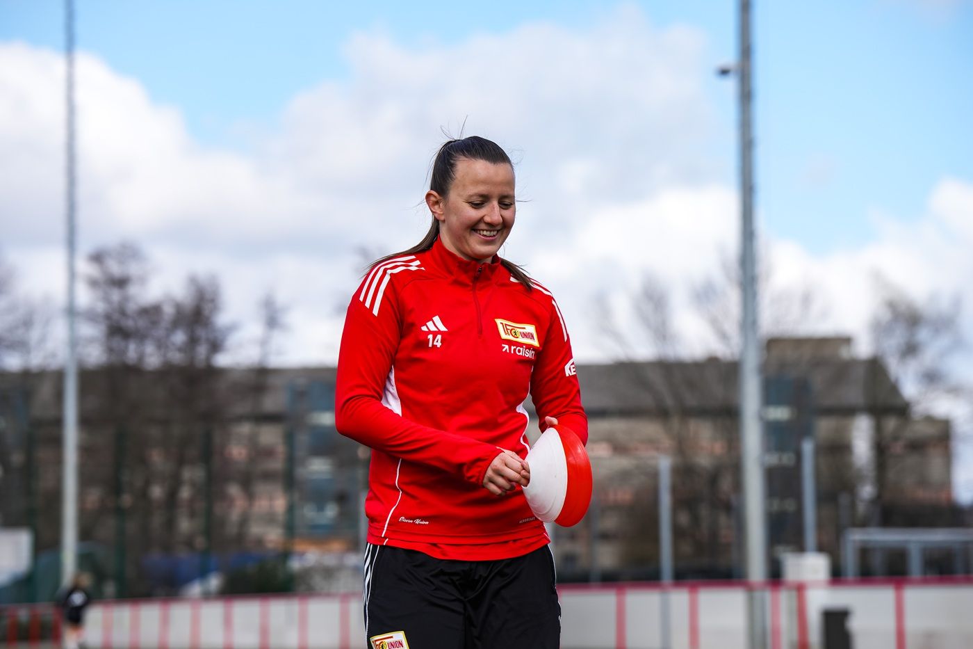 Eine sportlich gekleidete Frau mit längeren Haaren lächelt beim Training auf dem Fußballplatz.