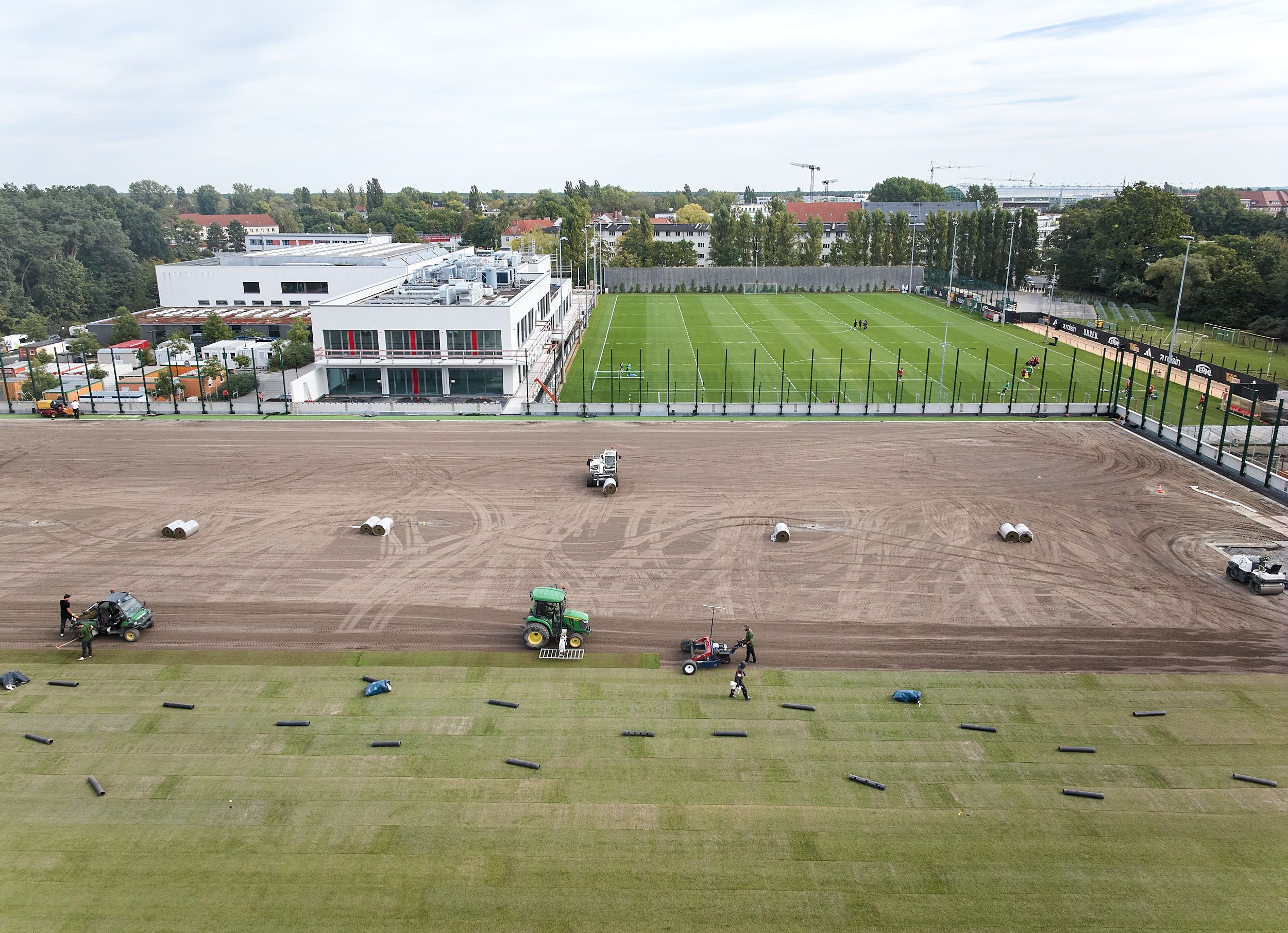 Blick auf einen Sportplatz mit frischem Rasen, Bauarbeiten und Maschinen zur Platzpflege im Vordergrund.