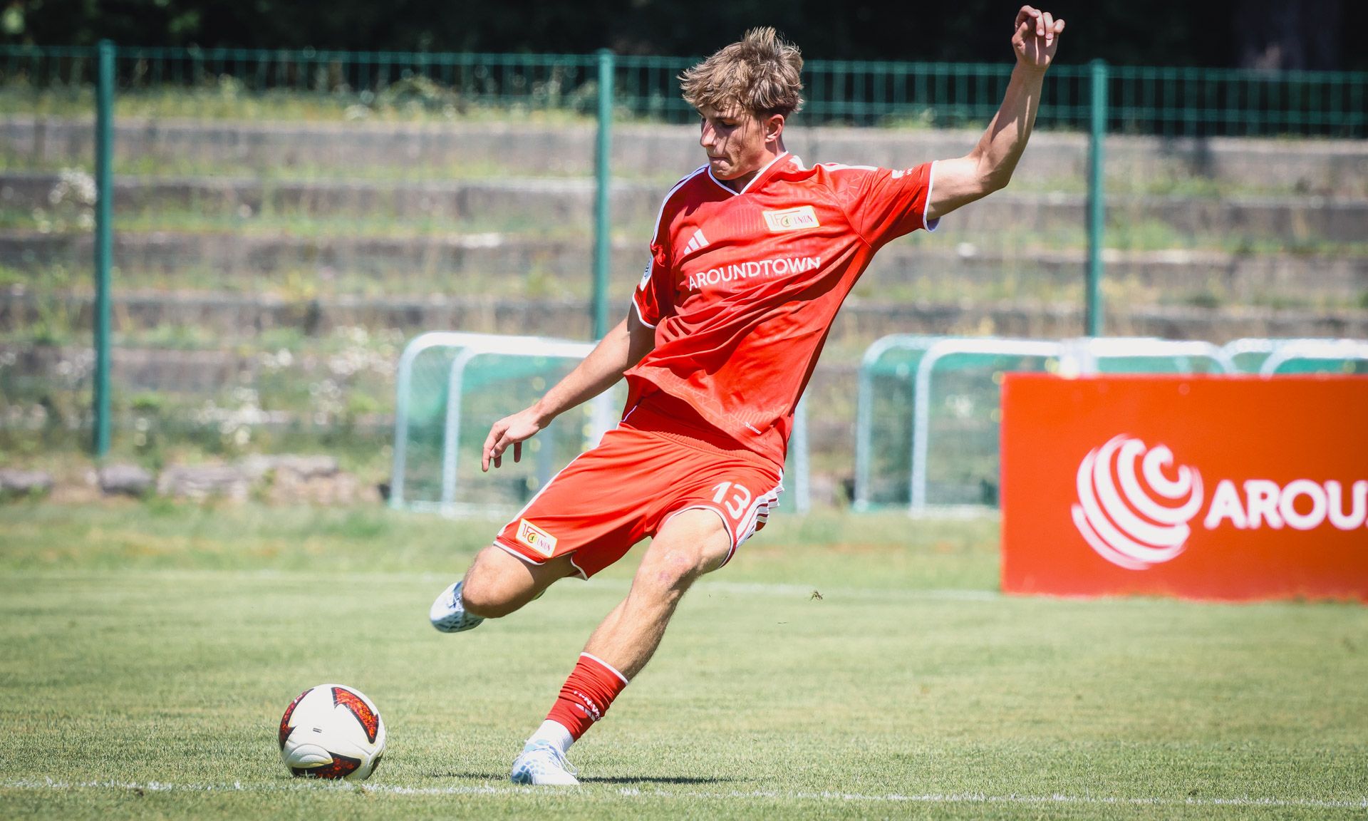 Ein Fußballspieler in einem roten Trikot führt einen Schuss auf das Tor aus, während er auf dem grünen Rasen spielt.