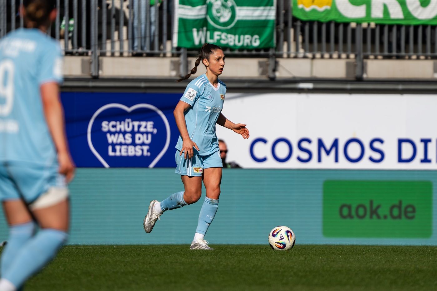 Eine Fußballspielerin in hellblauem Trikot dribbelt den Ball auf einem Spielfeld, im Hintergrund sind Fanbanner zu sehen.