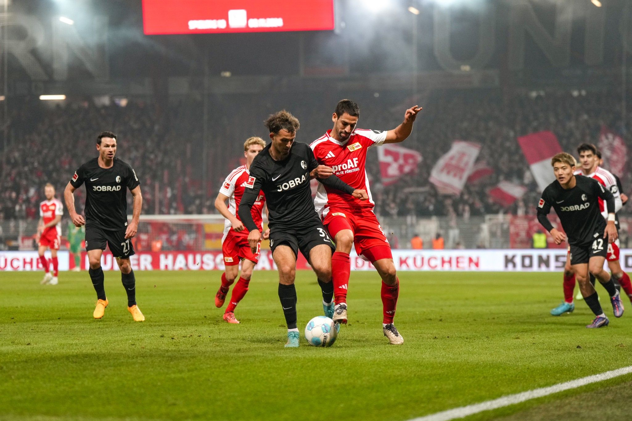 Ein Fußballspiel mit Spielern in schwarzen und roten Trikots, die um den Ball kämpfen, vor einer lebhaften Zuschauermenge.