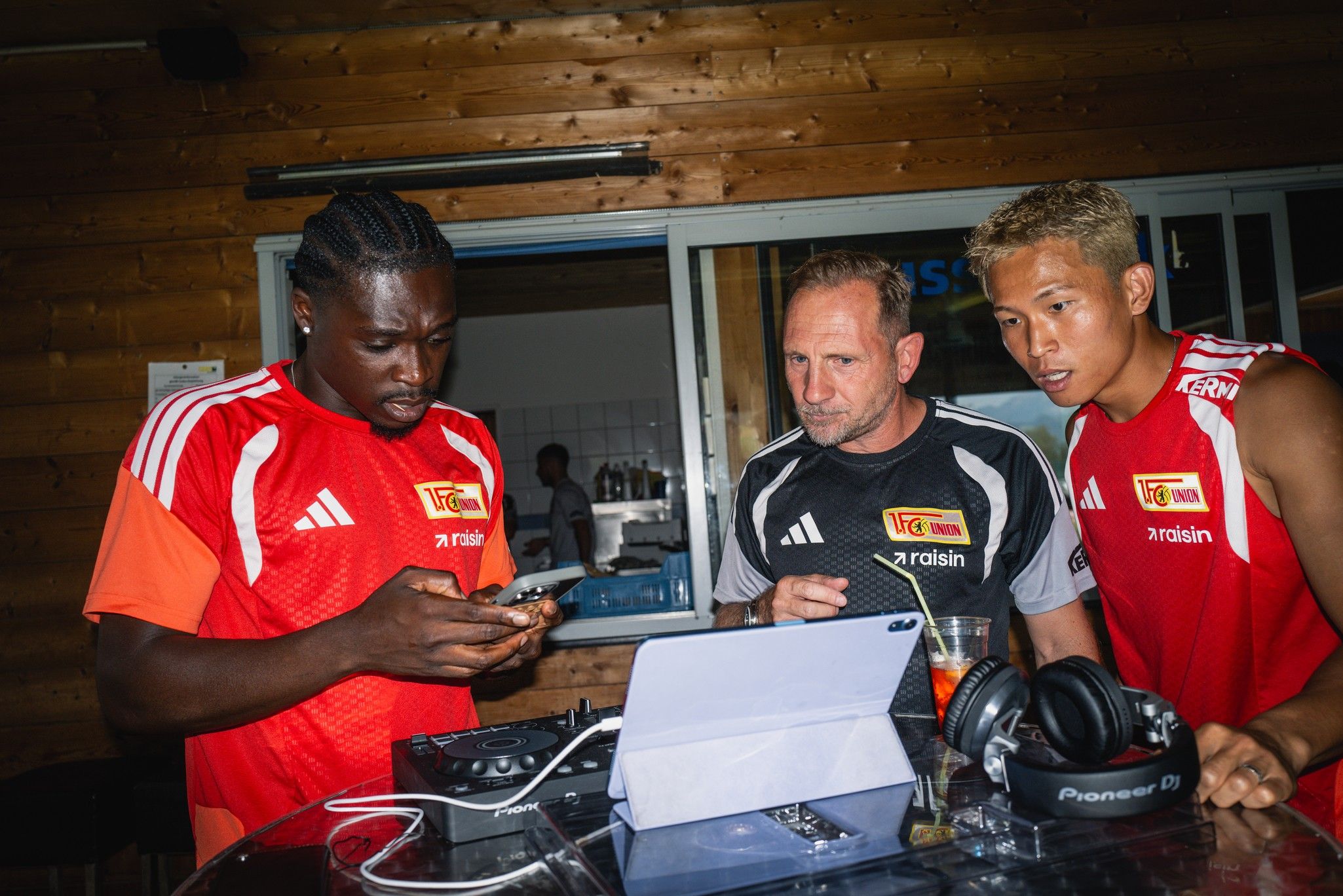 Drei Personen in roten Sporttrikots stehen in einem Raum, schauen auf ein Handy und stehen vor einer DJ-Ausrüstung.