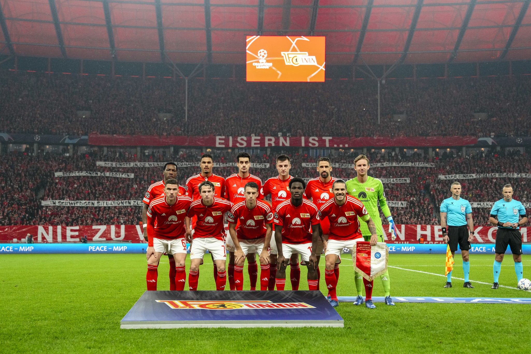 Eine Fußballmannschaft im rot-weißen Trikots steht auf dem Spielfeld vor unterstützendem Publikum und Banner.