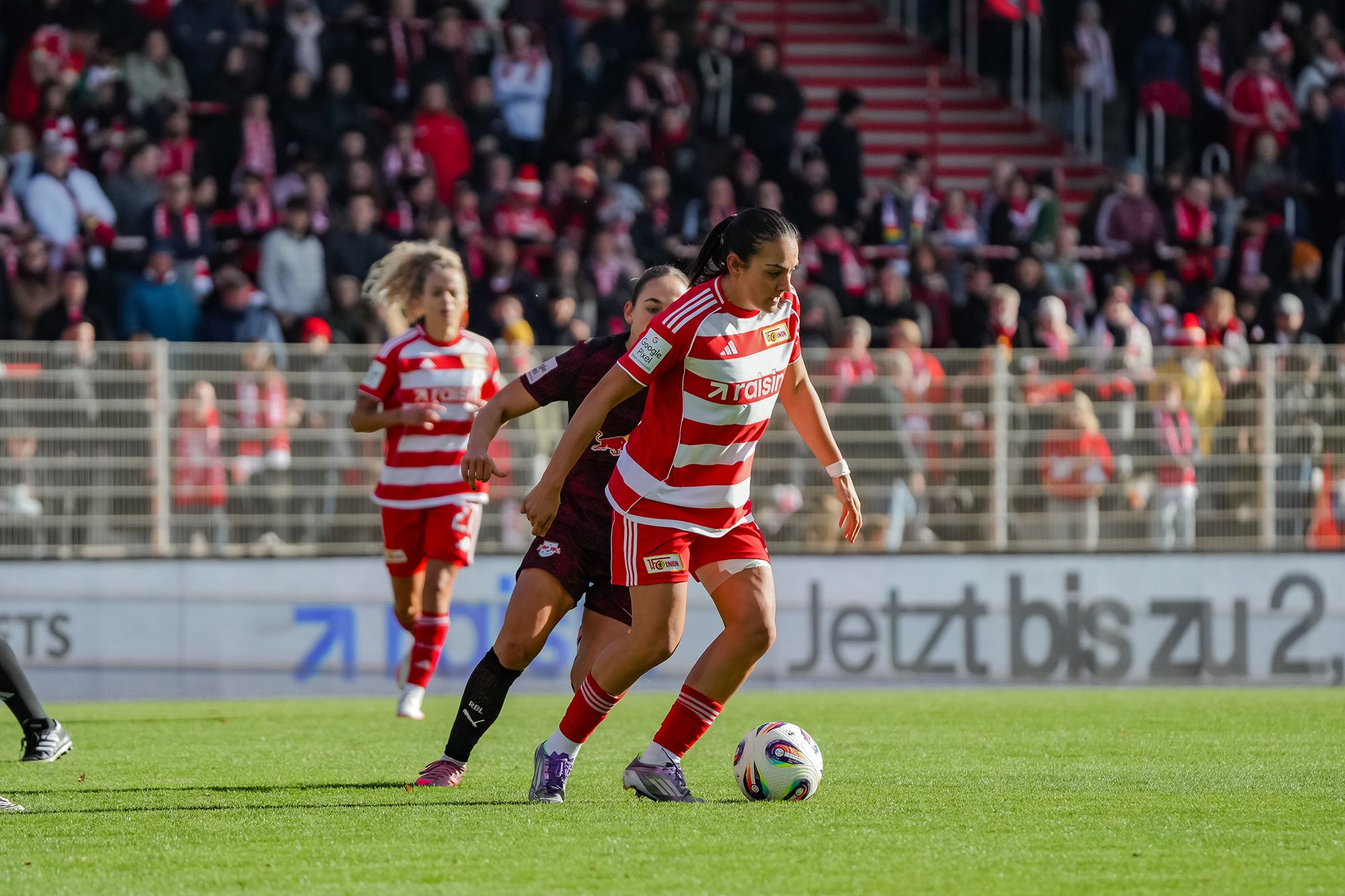 Eine Spielerin im rot-weißen Trikot dribbelt den Ball auf dem Spielfeld, während eine Gegenspielerin ihr nahekommt. Zuschauer im Hintergrund.