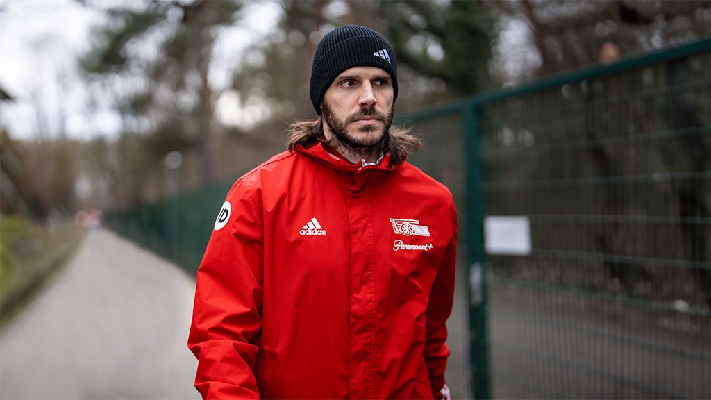 Man in a red tracksuit with a logo and black hat walking along a path by a fence.