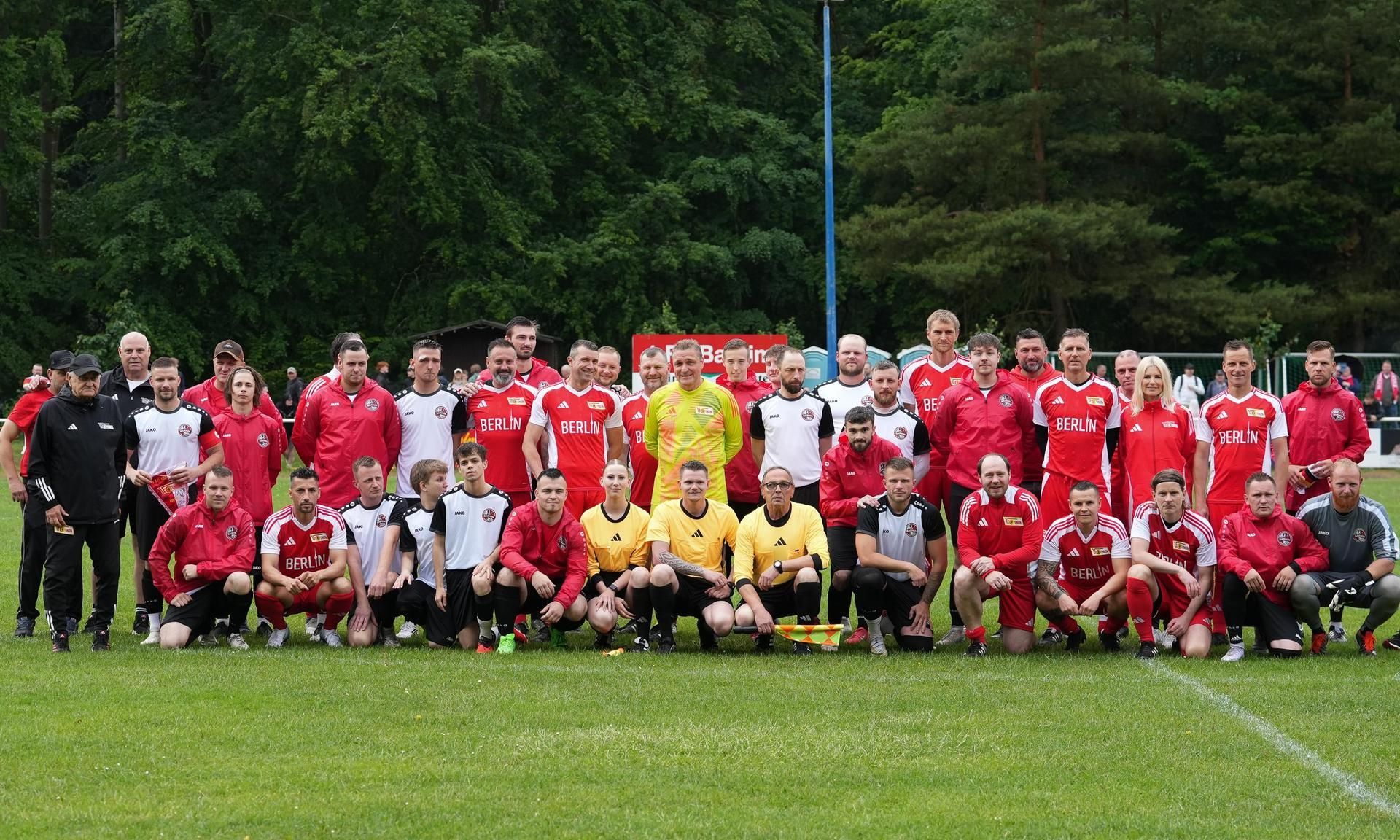Gruppenfoto von Fußballspielern in roten und schwarzen Trikots auf einem Rasenfeld, umgeben von Bäumen.