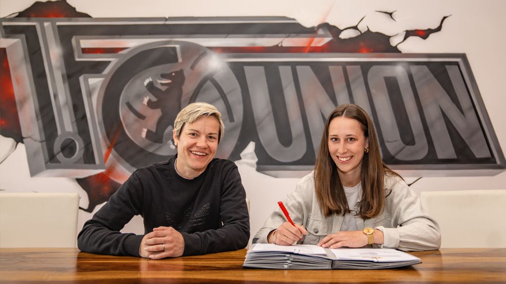 Zwei Frauen sitzen an einem Tisch, lächeln und posieren vor einer Wand mit dem Logo von Union. Eine von ihnen unterschreibt ein Dokument.