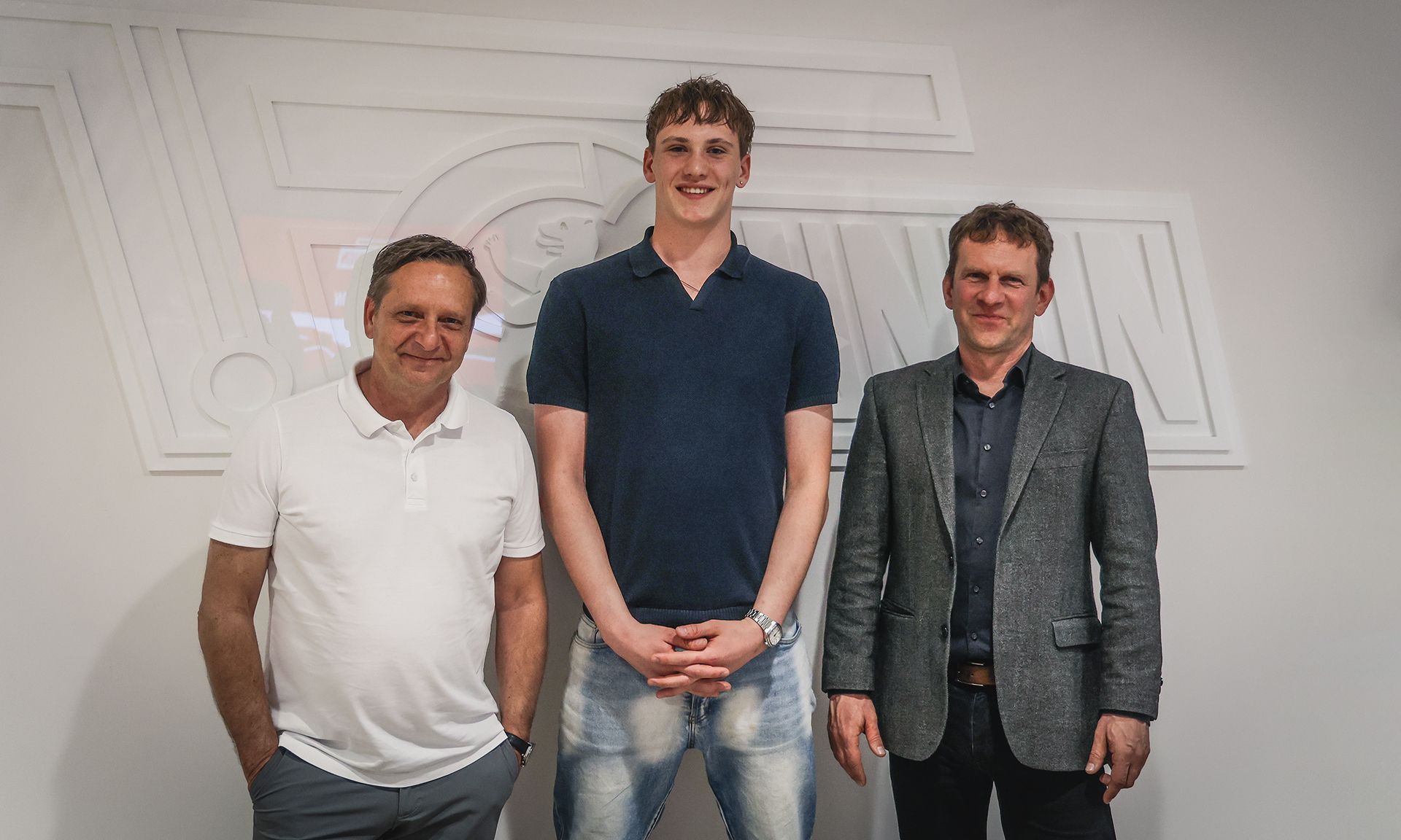Drei Personen stehen vor einer hellen Wand mit einem Relief. Die erste Person trägt ein weißes Shirt, die zweite ein blaues Polo und die dritte einen grauen Anzug.