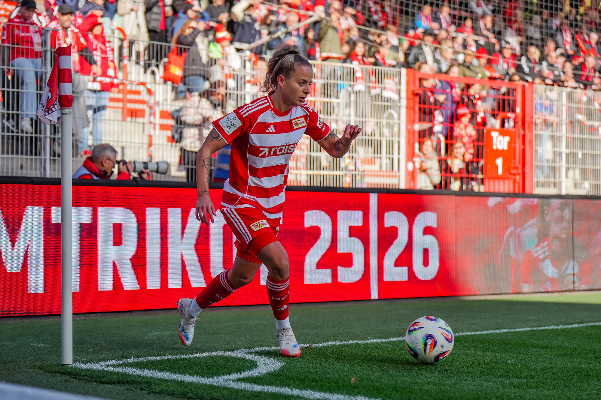 Eine Fußballspielerin in rot-weißem Trikot führt einen Eckball aus, während Zuschauer im Hintergrund jubeln.