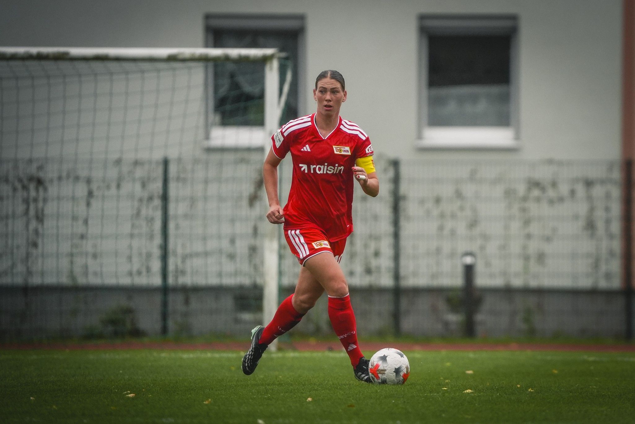 Eine fußballspielende Frau in roten Trikots mit einer Kapitänsbinde sprintet über das Spielfeld.