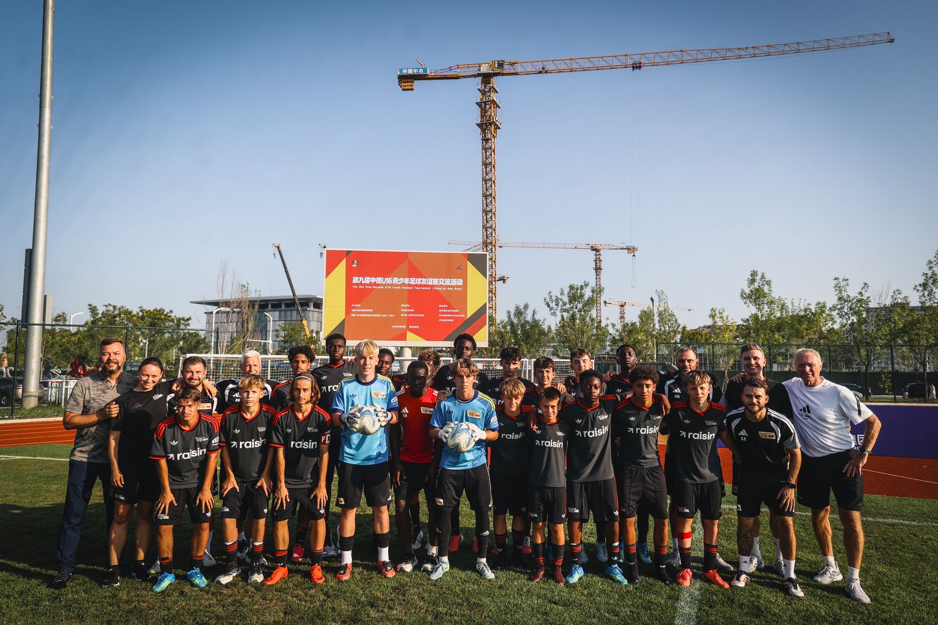 Gruppe von Fußballspielern und Trainern posiert auf einem Platz, im Hintergrund ist ein Baukran und ein Infotunnel zu sehen.