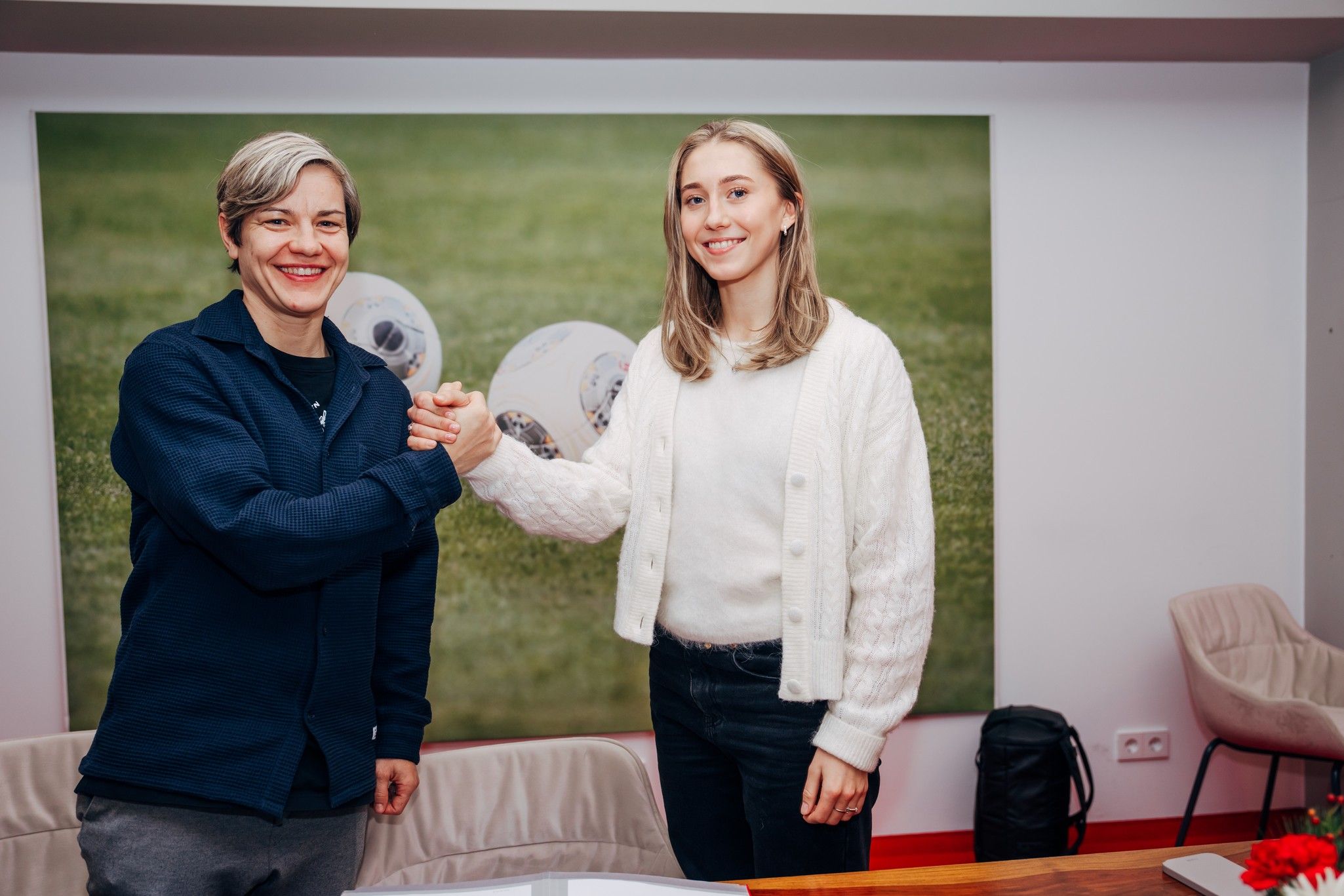 Zwei Frauen schütteln sich die Hände in einem Raum, im Hintergrund sind Fußball-Dekorationen zu sehen.