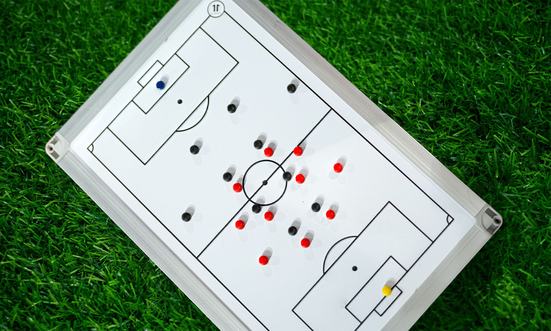 Ein Fußball-Taktikboard mit rot, schwarz und blau markierten Spielfiguren auf grünem Kunstrasen.
