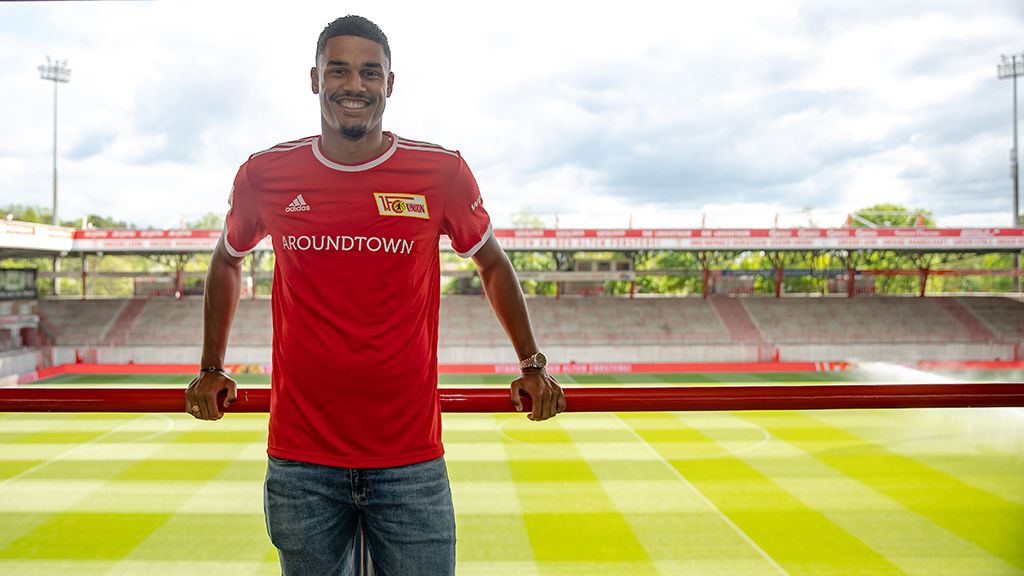 Ein junger Mann in einem roten Fußballtrikot steht lächelnd an einem Geländer in einem Stadion mit Rasenfeld im Hintergrund.