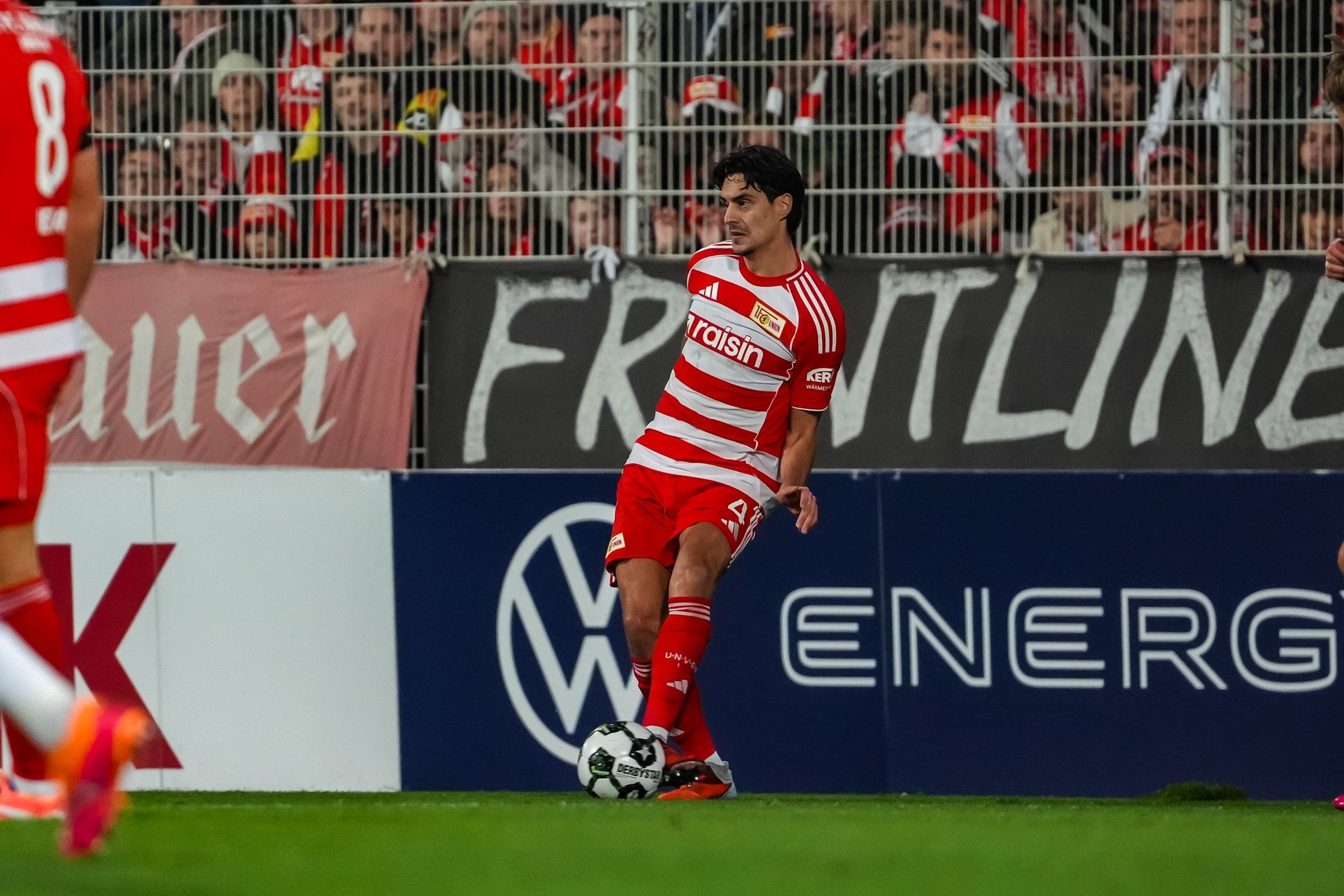 Ein Fußballspieler in roter Vereinsausrüstung führt einen Einwurf aus, während Zuschauer im Hintergrund zu sehen sind.
