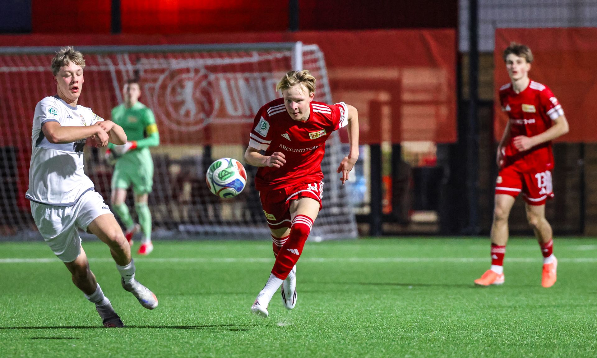 Ein Fußballspieler in rotem Trikot sprintet mit dem Ball, während ein Gegner in Weiß ihn verfolgt. Im Hintergrund ist ein Tor zu sehen.
