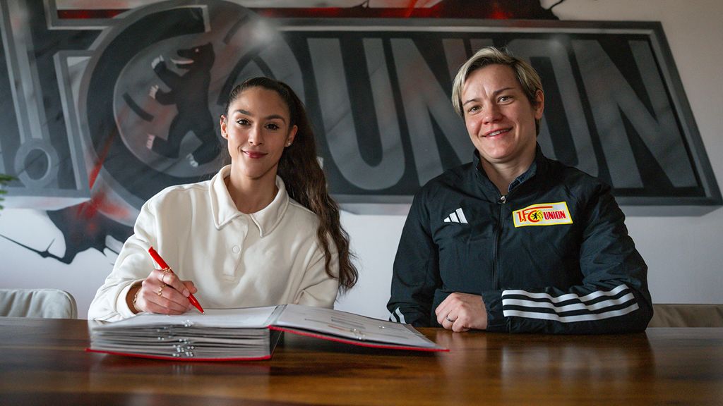 Two women sit at a table signing documents, with a club logo in the background.
