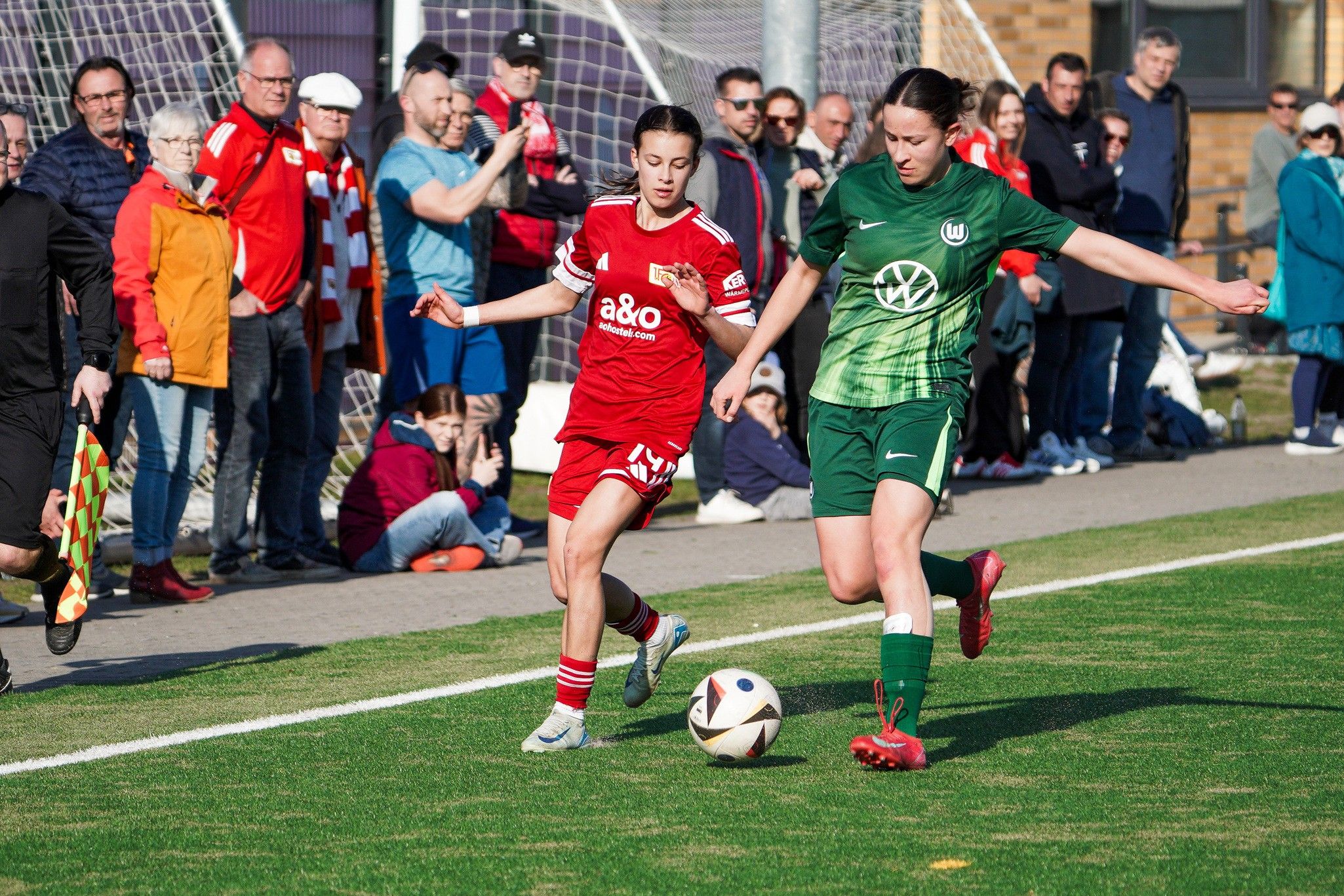 Eine Fußballspiel-Szene mit zwei Spielerinnen, einer in Rot und einer in Grün, umgeben von Zuschauern.
