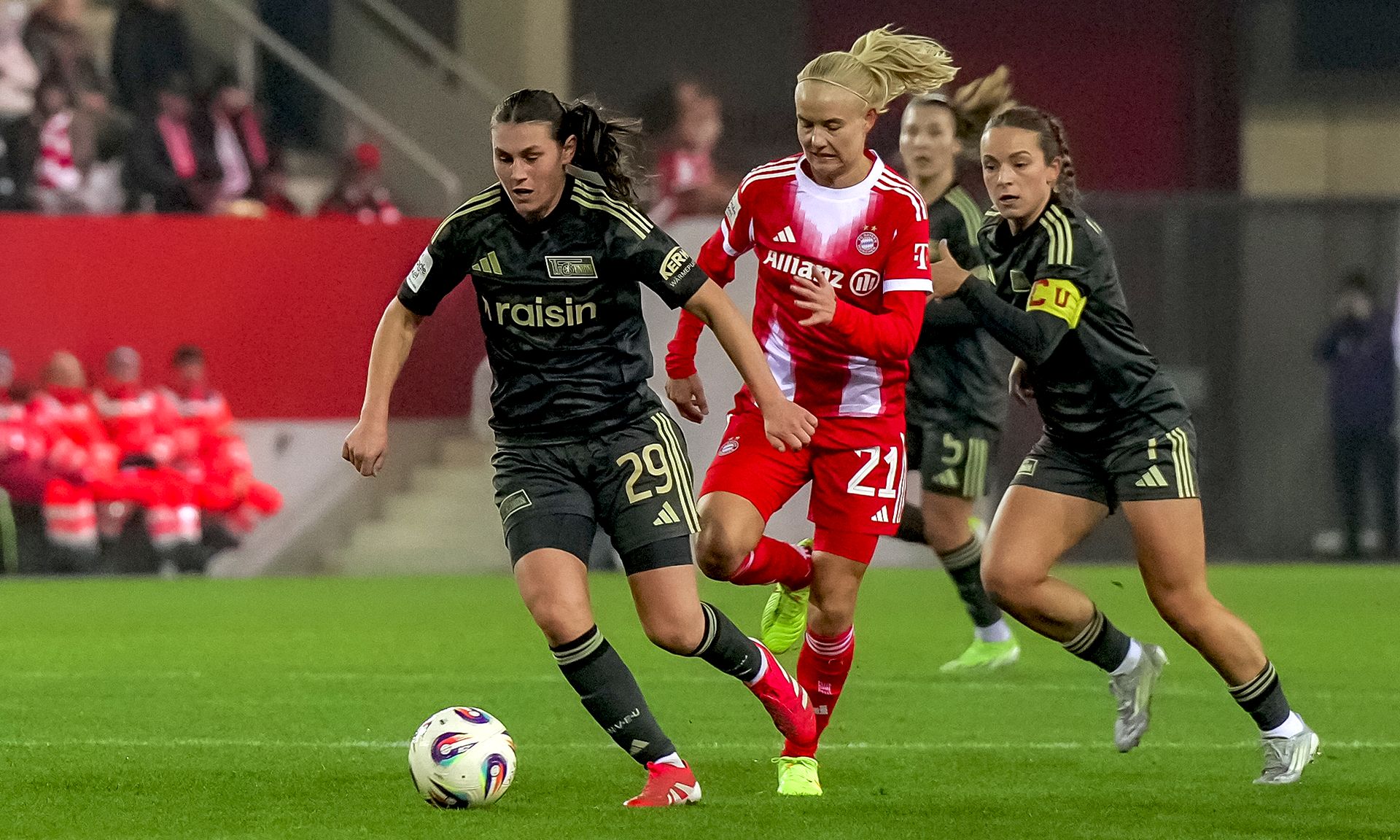 Eine Fußballspielszene mit zwei Spielerinnen in schwarzen Trikots und einer in einem roten Trikot, die um den Ball kämpfen.