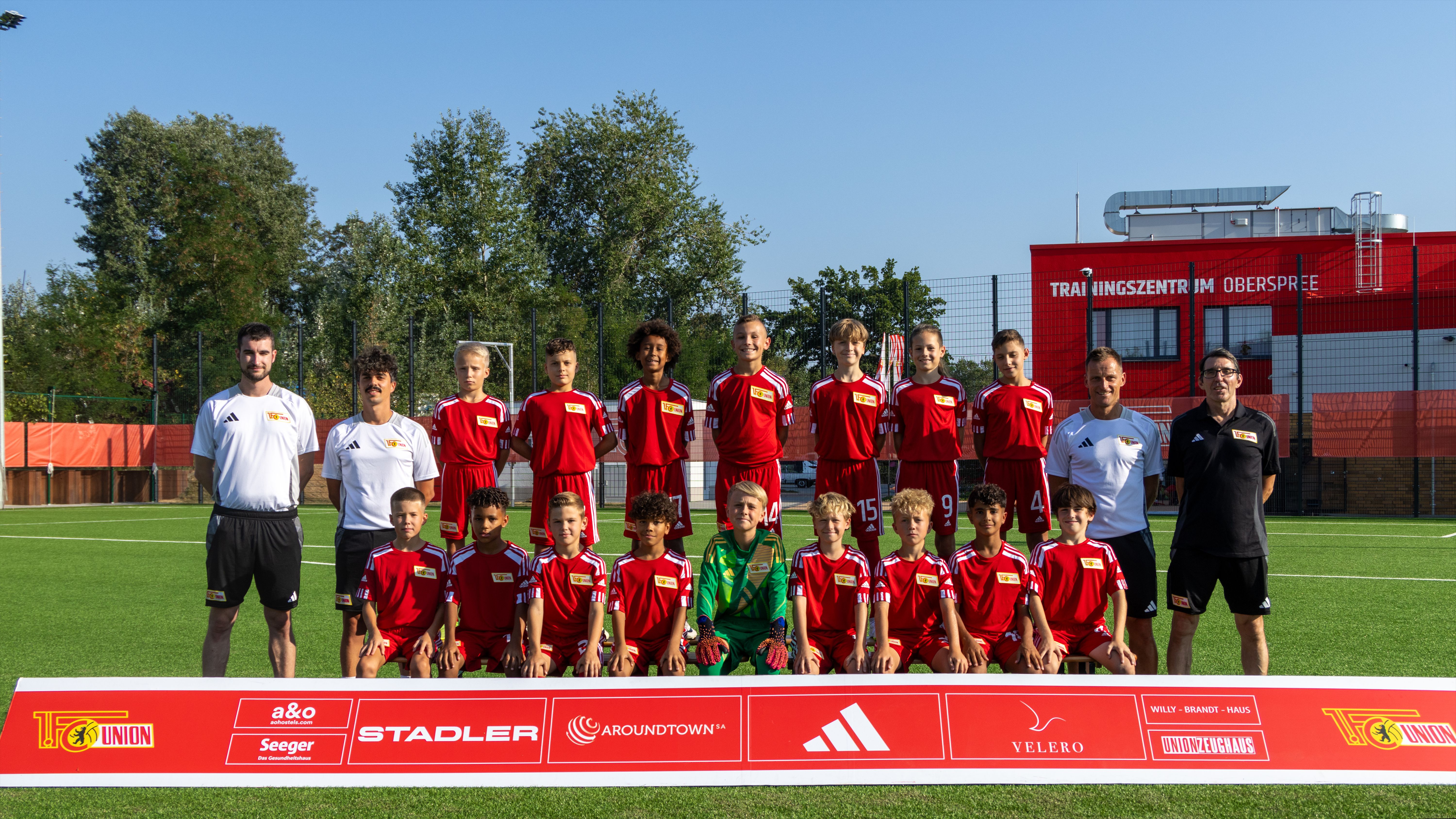 Eine Fußballmannschaft in roten Trikots posiert auf einem Platz. Im Hintergrund sind Bäume und ein Gebäude zu sehen.