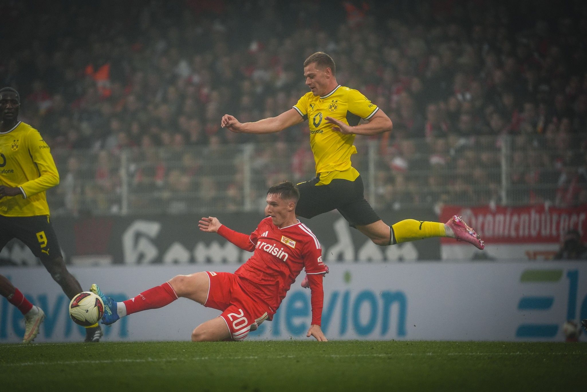 Ein Fußballspieler im roten Trikot rutscht auf dem Platz, während ein Spieler im gelben Trikot über ihn springt.