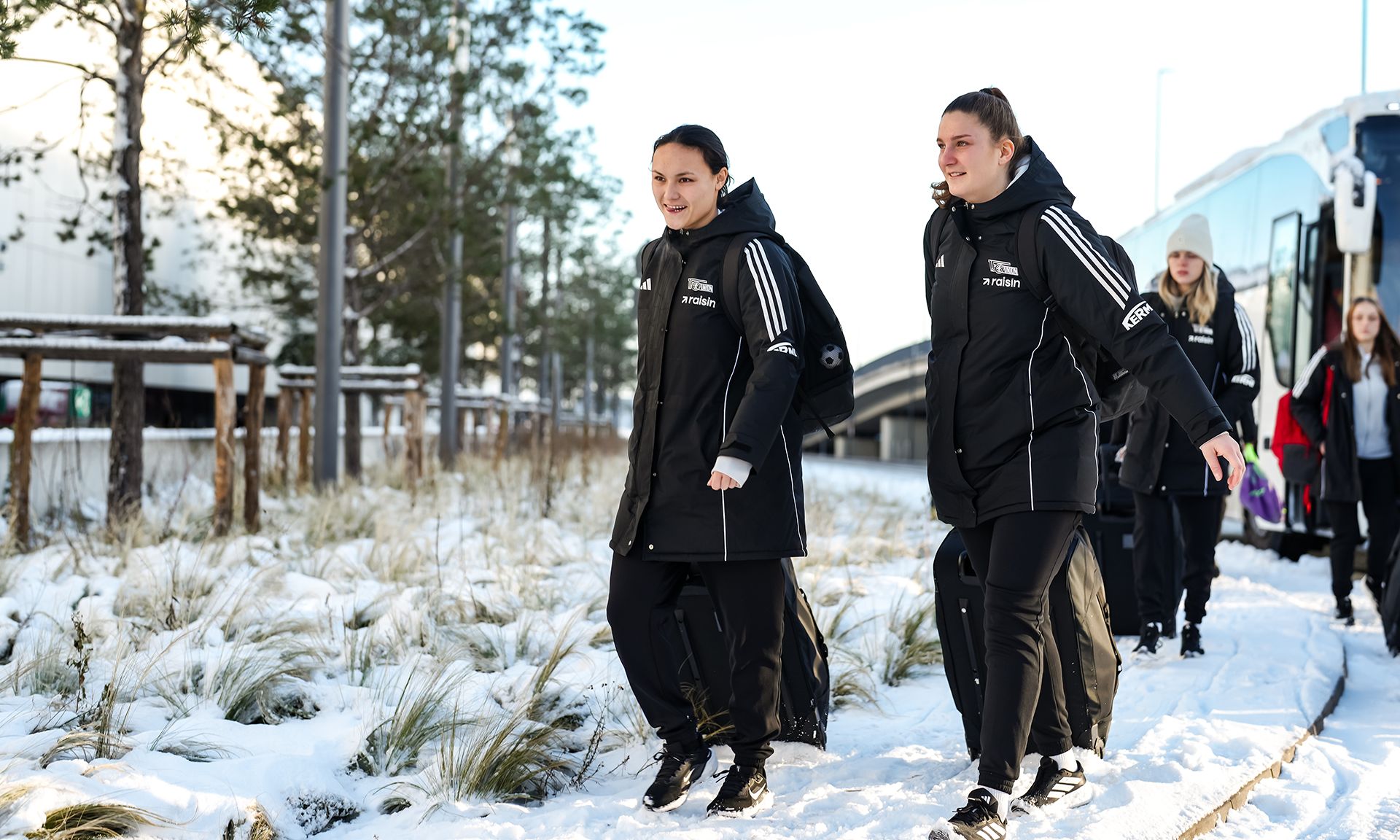 Zwei Personen in Winterjacken gehen auf einem schneebedeckten Weg vorbei an Bäumen und einem Bus.