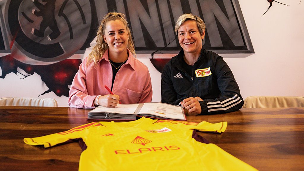 Zwei Frauen sitzen an einem Tisch, unterzeichnen Dokumente und zeigen ein Fußballtrikot in Gelb mit dem Aufdruck „ELARIS“.