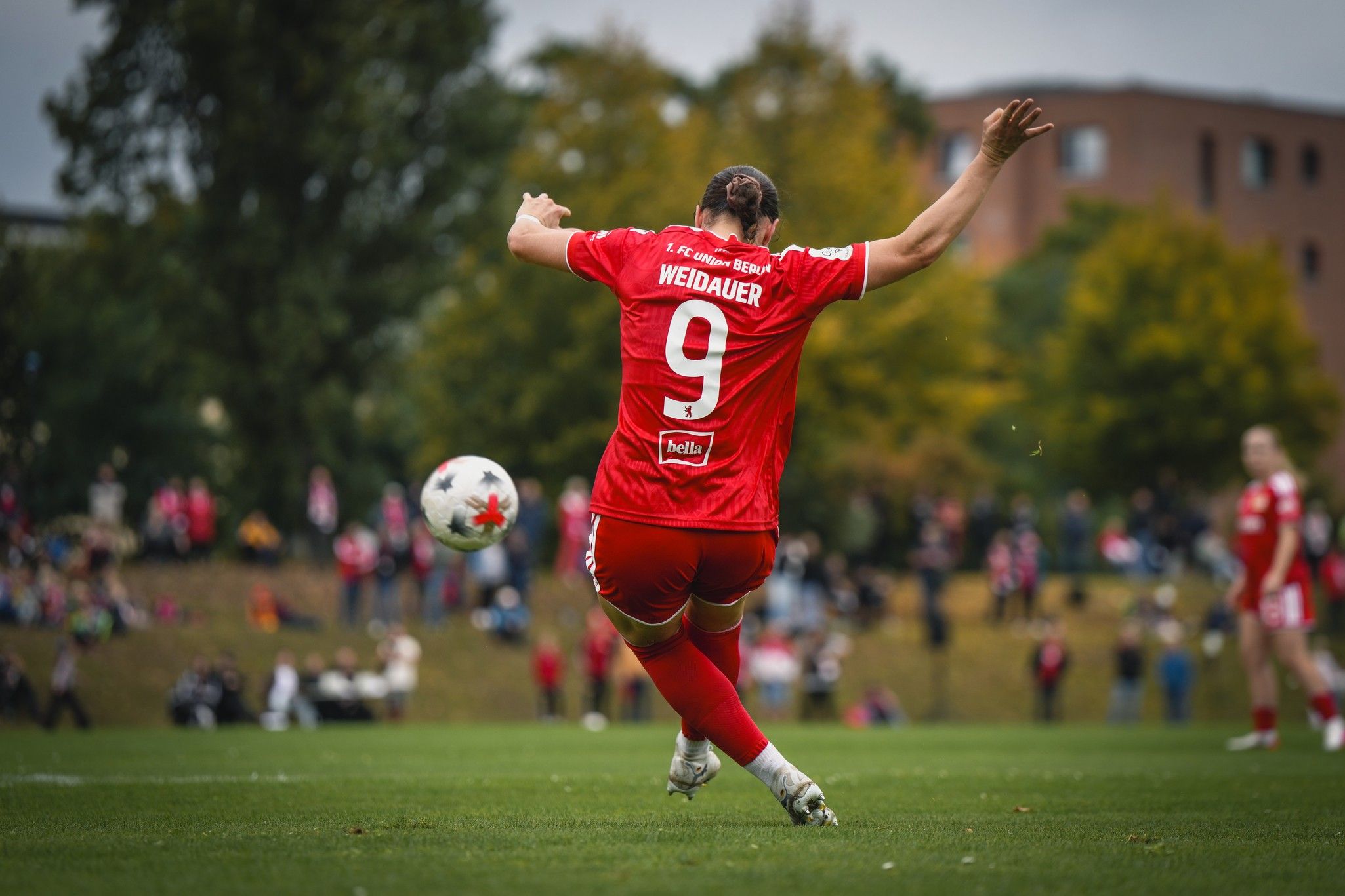 Spieler mit der Nummer 9 in rotem Trikot schießt den Ball auf einem Rasenplatz, umgeben von Zuschauern und Bäumen.