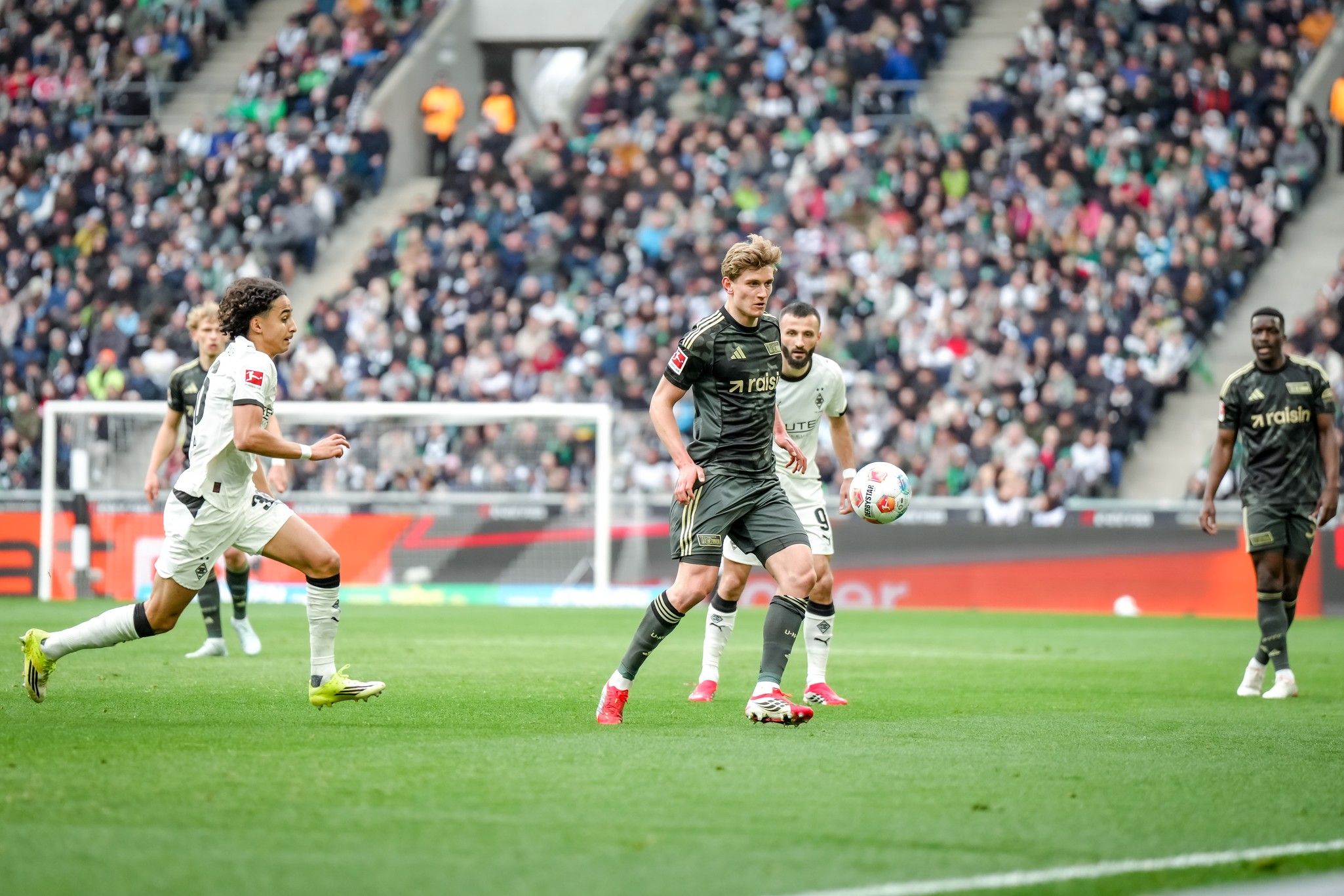 Spielszene in einem Stadion: Ein Spieler passt den Ball, während zwei Gegner im Hintergrund darauf reagieren. Fans sind im Stadion zu sehen.