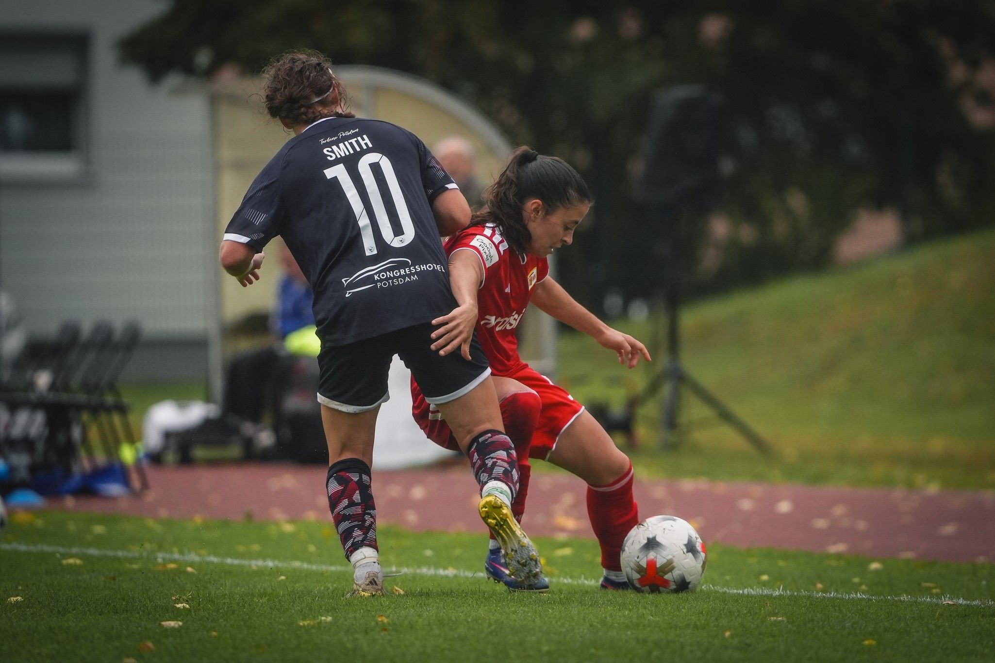 Zwei Fußballspielerinnen im Spiel; eine Spielerin in roten Trikots dribbelt, während die andere in schwarzem Trikot folgt.