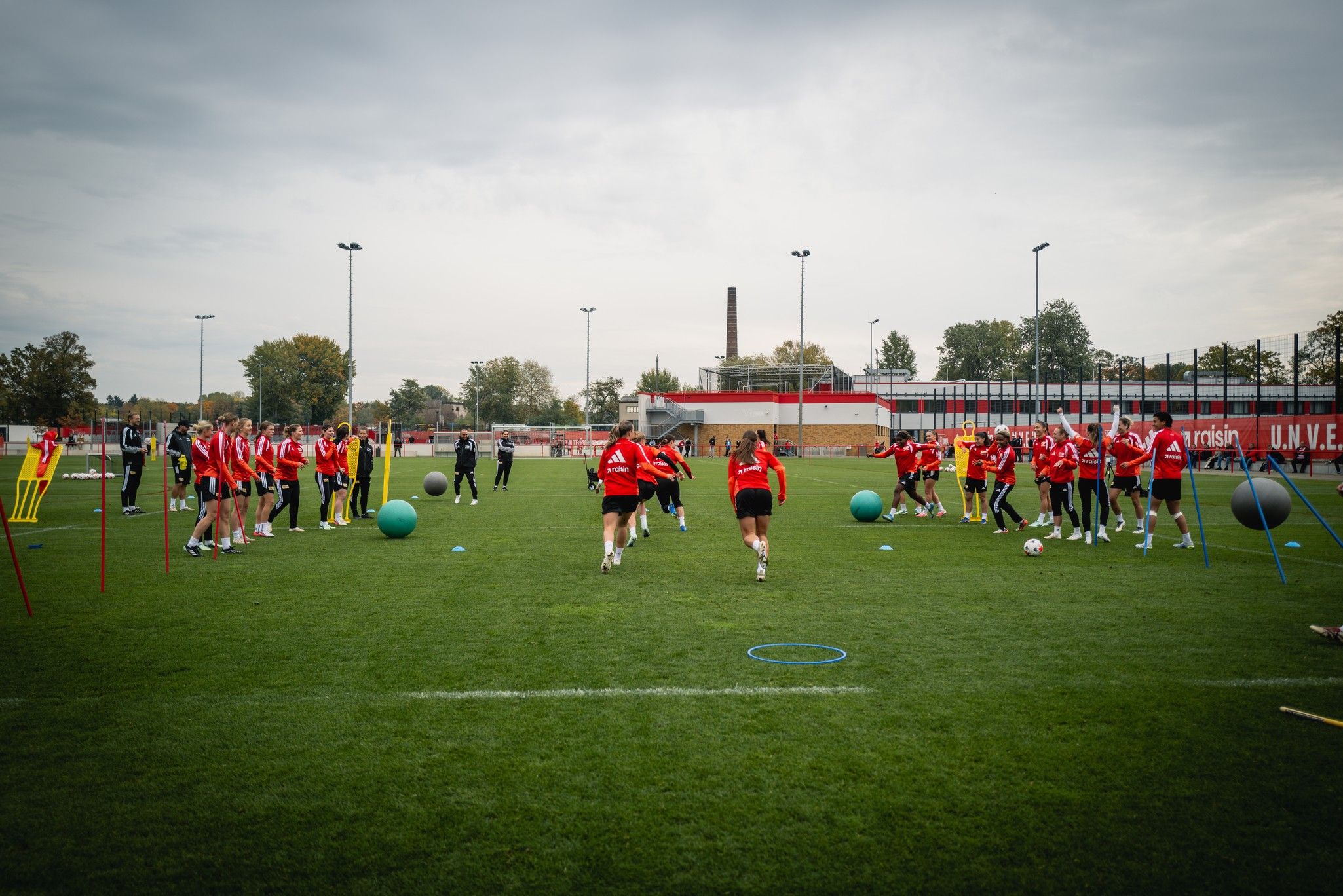 Fußballtraining auf einem Rasenplatz mit Spielern in roten Trikots, Übungen mit Bällen und Hindernissen.