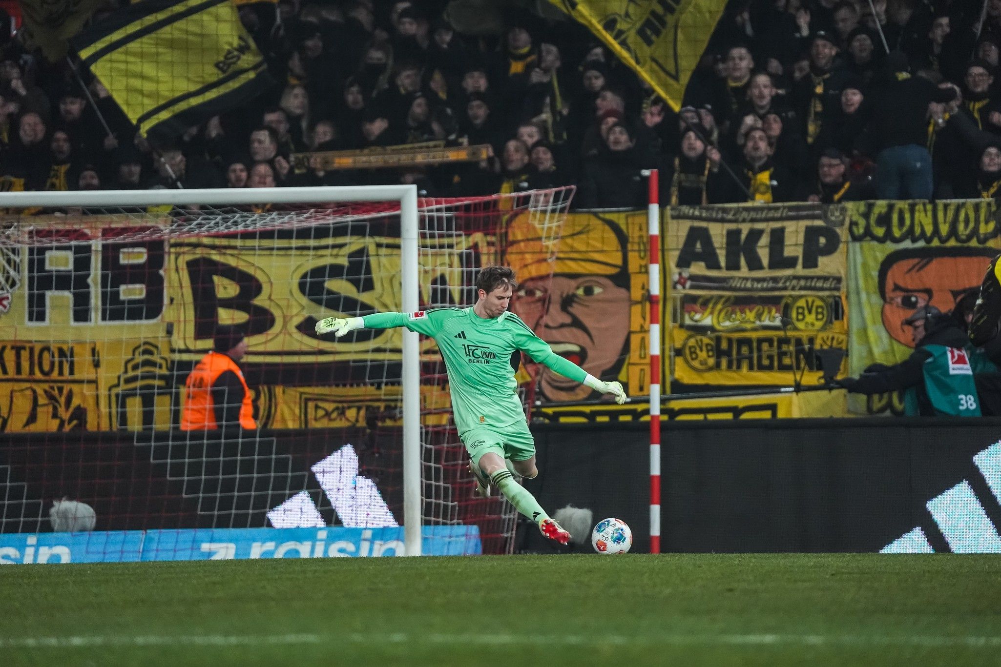 Ein Torwart in grüner Ausrüstung steht bereit, während Fans mit gelben und schwarzen Fahnen im Hintergrund jubeln.