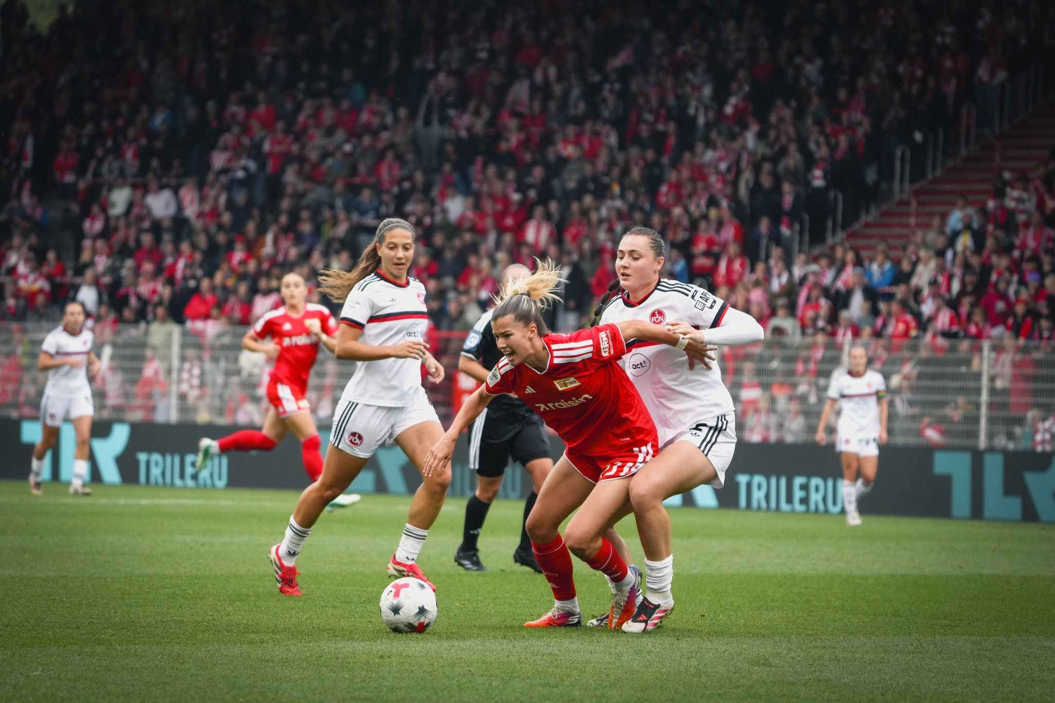 Eine Fußballspielszene mit zwei Spielerinnen im roten Trikot, die um den Ball kämpfen, während Zuschauer im Hintergrund stehen.